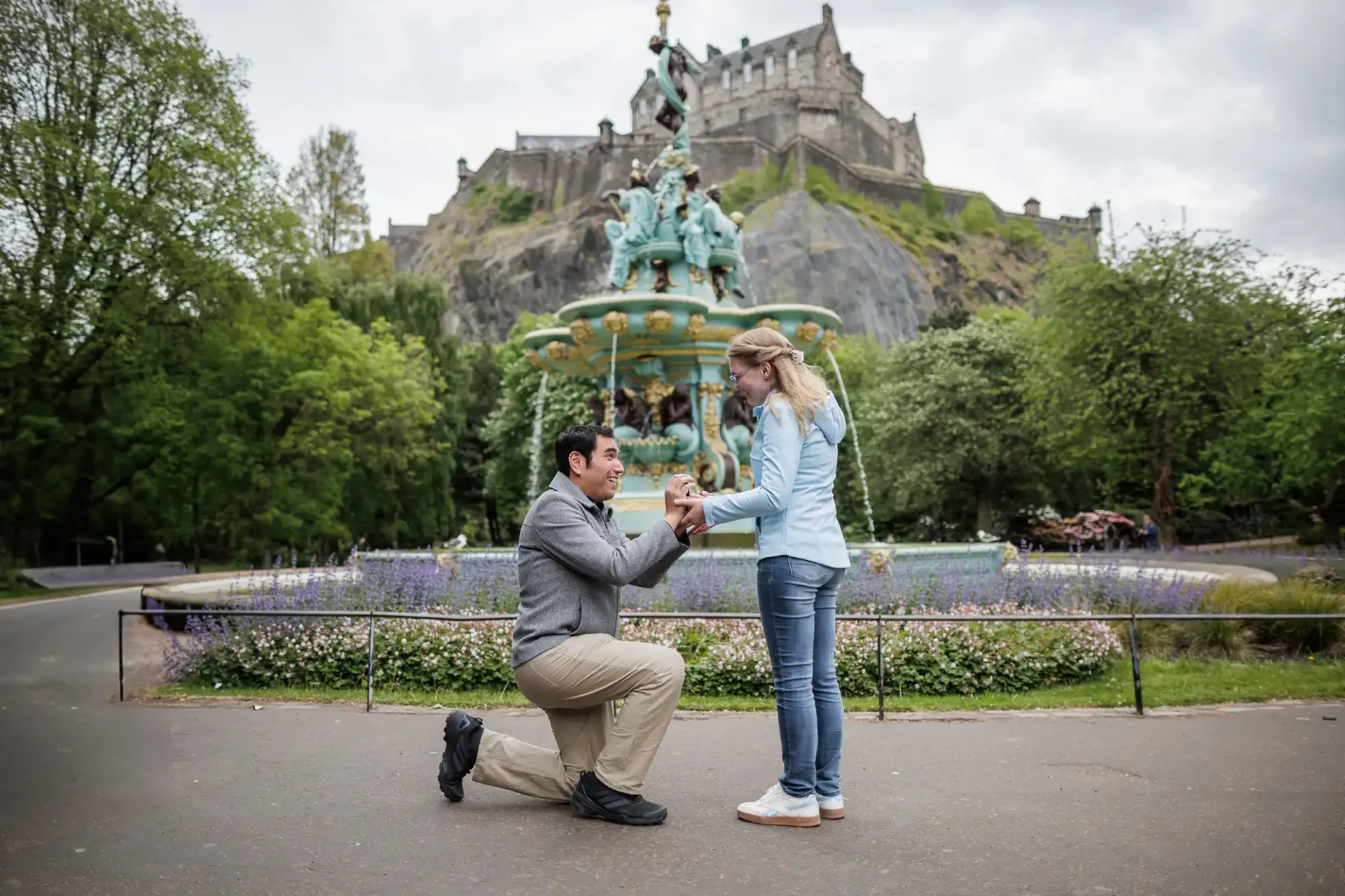 A man kneels to propose to a woman in front of a decorative fountain, with a historic castle on a hill in the background.