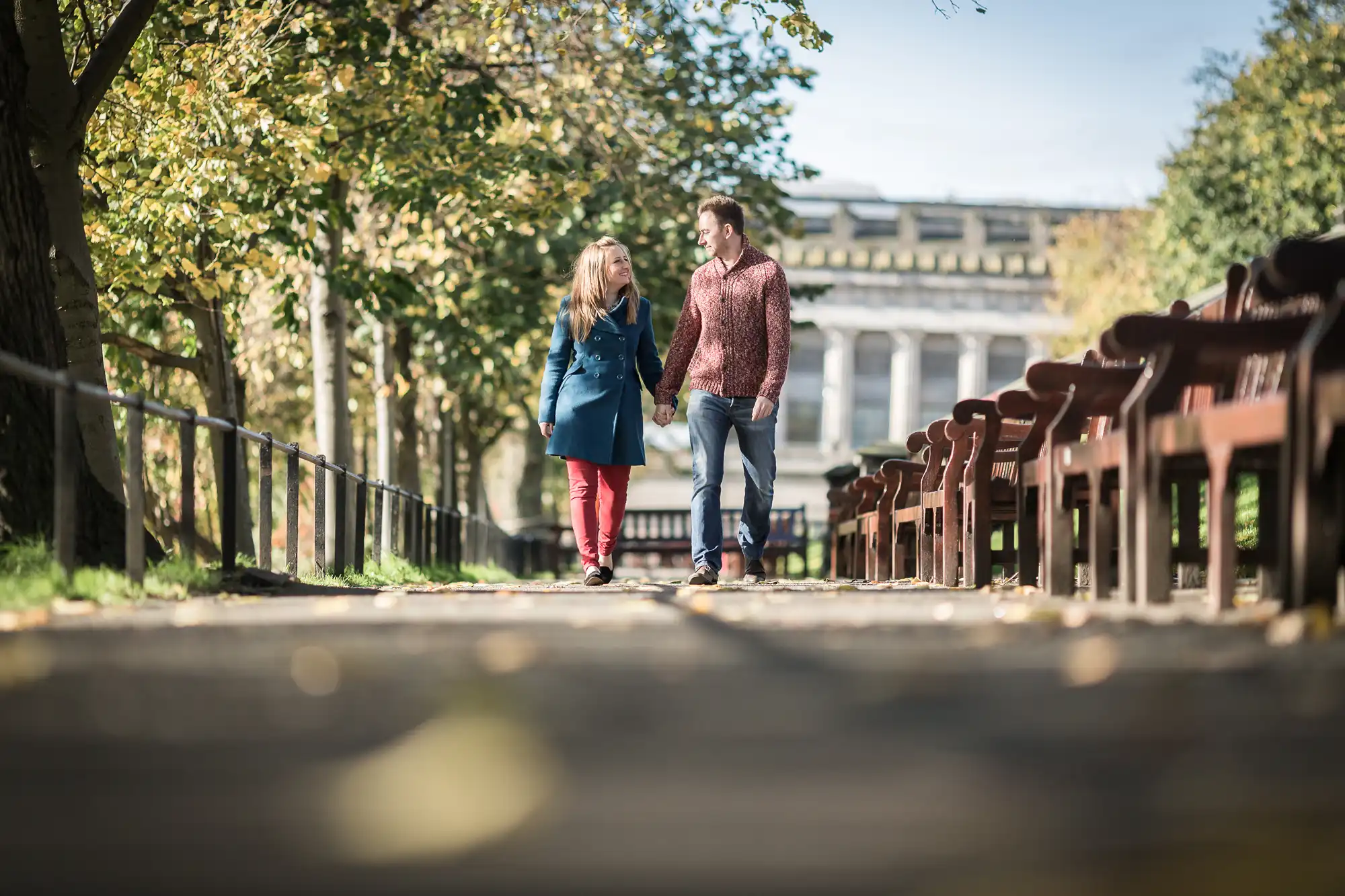 A couple walks hand in hand down a leafy park path, flanked by wooden benches, with classical architecture in the background on a sunny day.