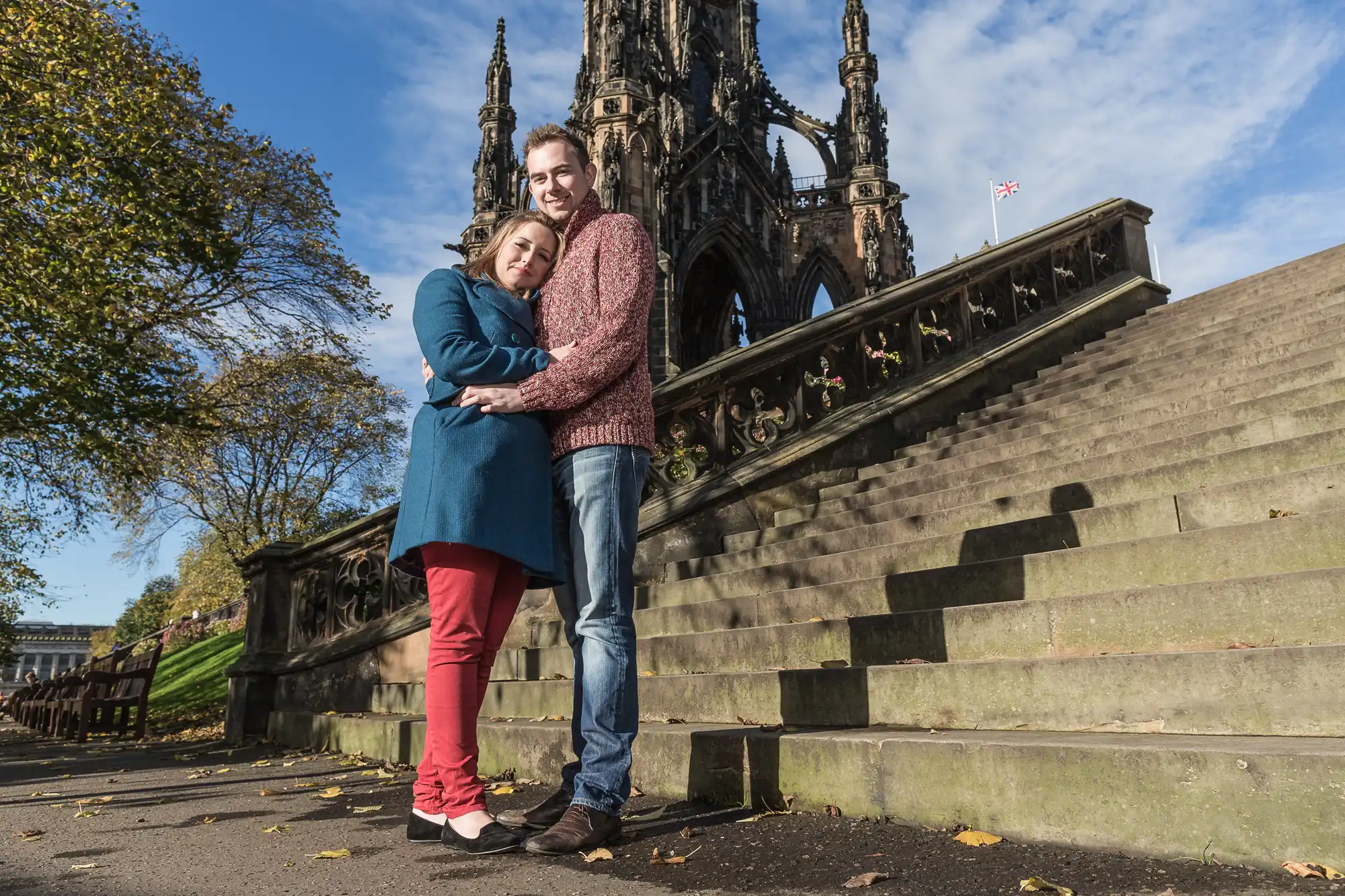 A couple embracing on the steps of a park with the intricate scott monument in the background, under a clear blue sky.