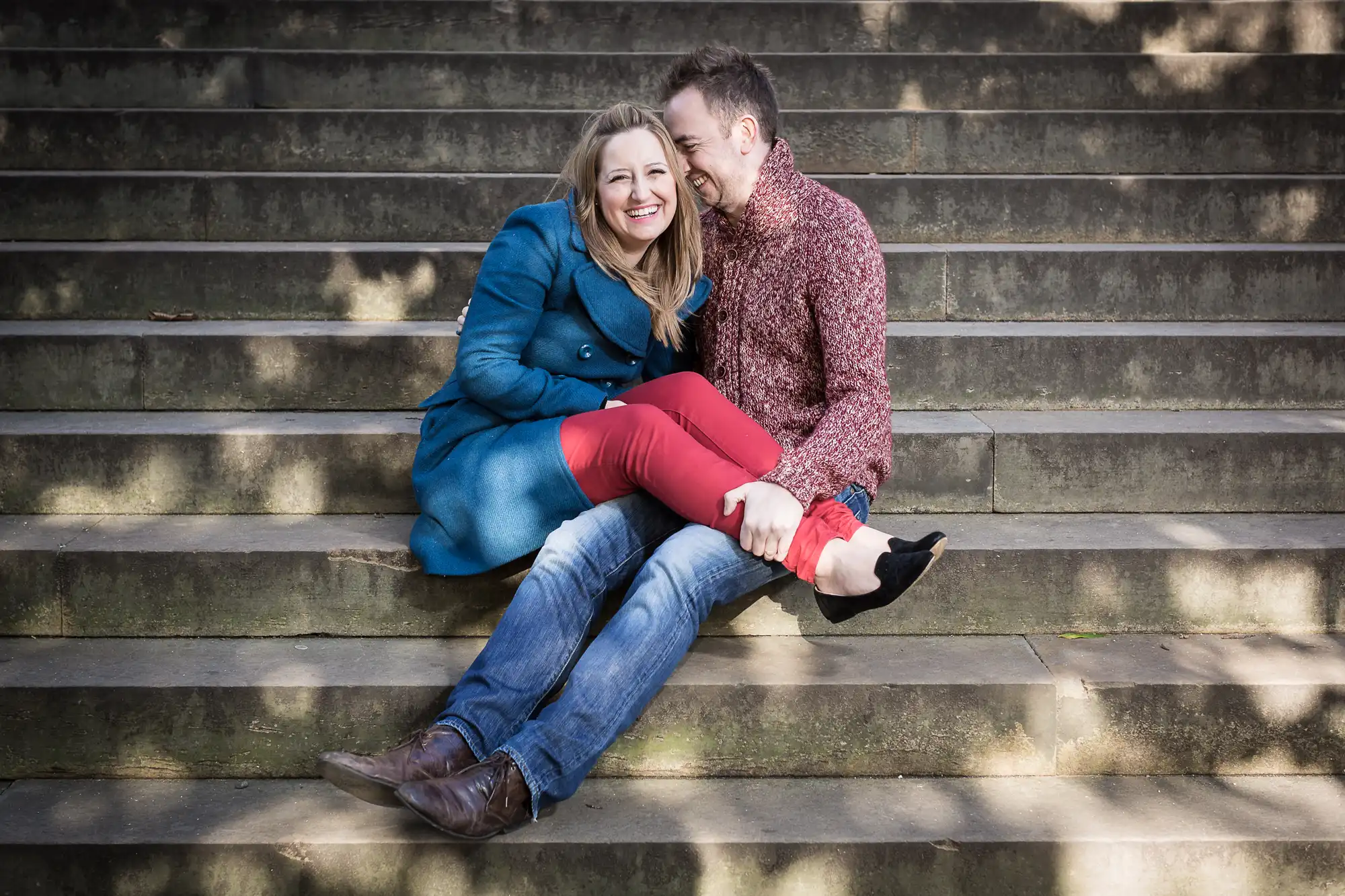 A joyful couple sitting closely and laughing on stone steps, with the man holding a woman's bare foot. she is wearing a blue coat and red pants; he is in a patterned shirt and jeans.