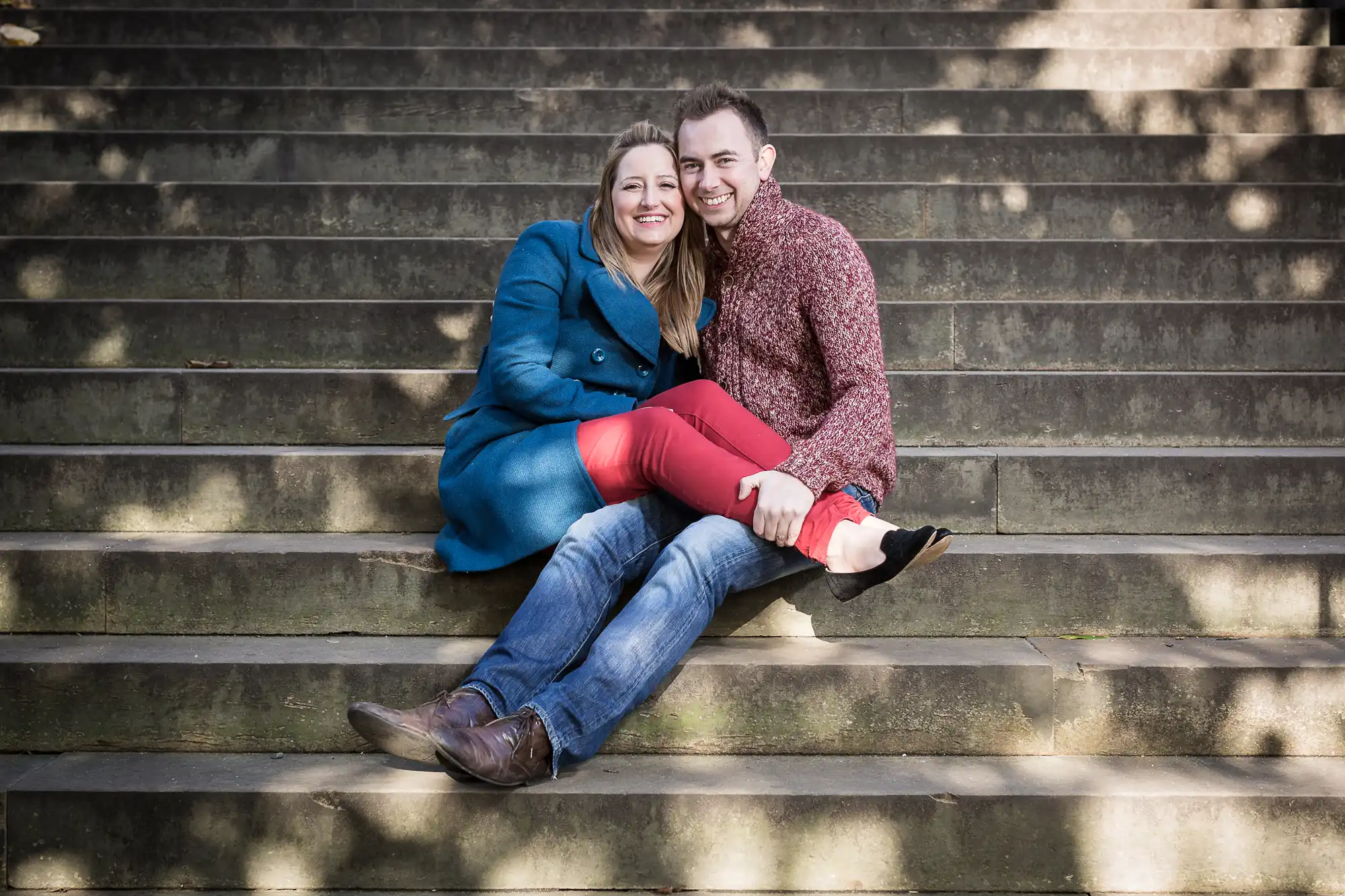 A happy couple sitting closely on stone steps, smiling at the camera, with the woman wearing a blue coat and the man in a red shirt and jeans.