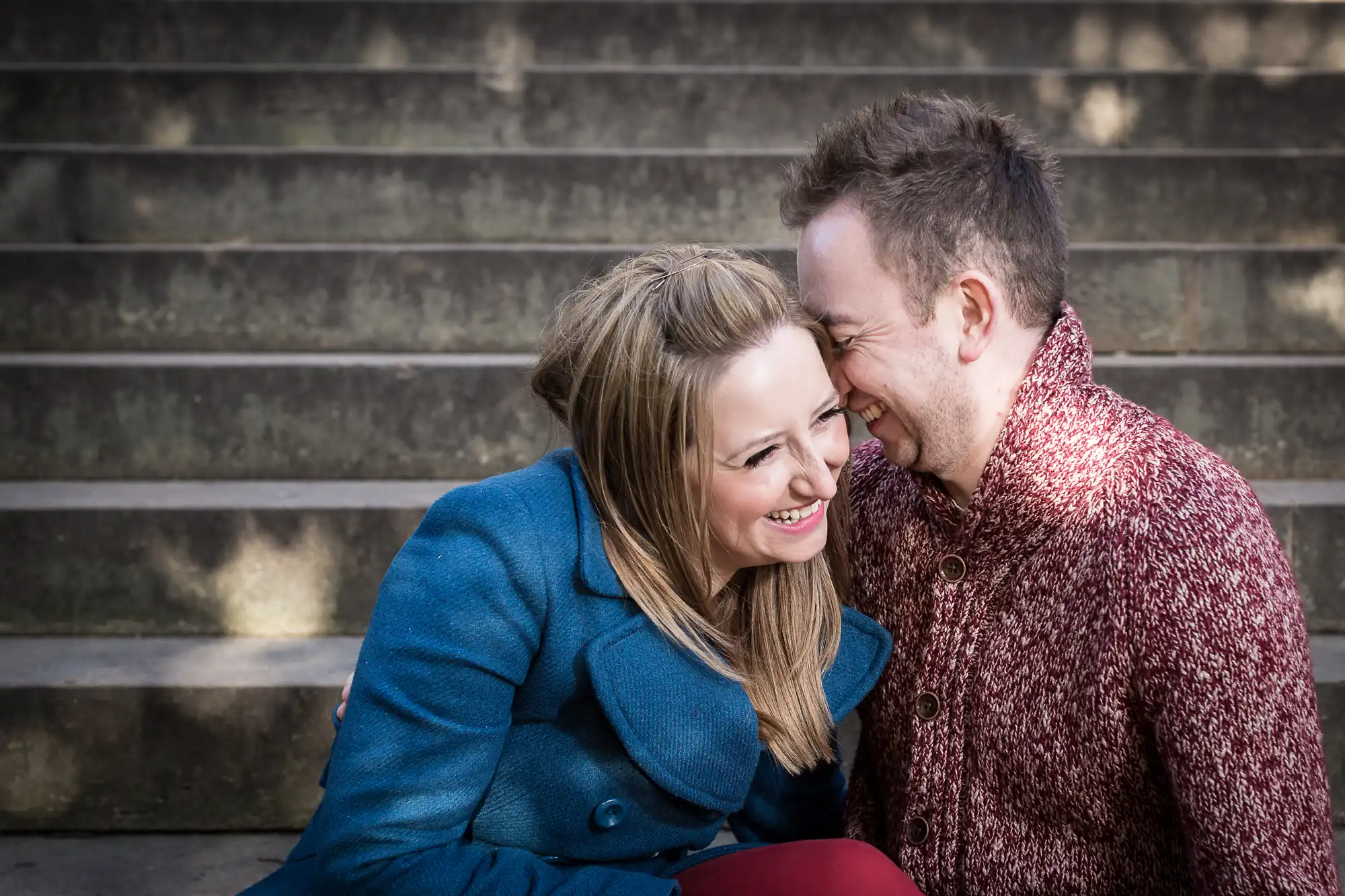 A joyful couple sitting closely and laughing together on outdoor steps. both are dressed in warm, casual clothing.