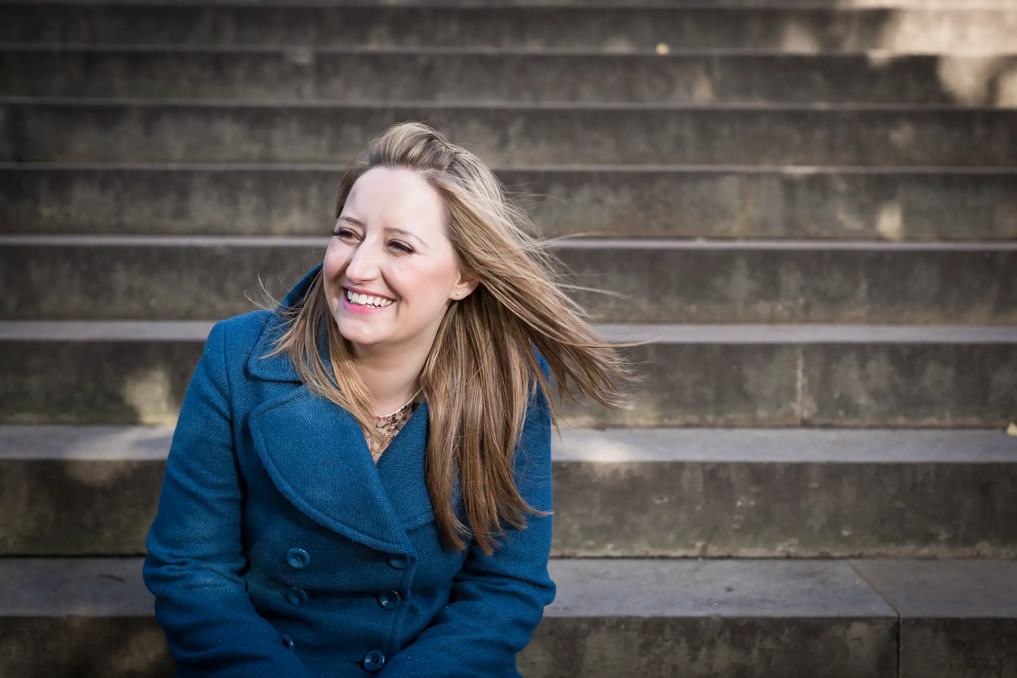 A woman in a blue coat laughing while sitting on outdoor steps. her hair blows slightly in the breeze.
