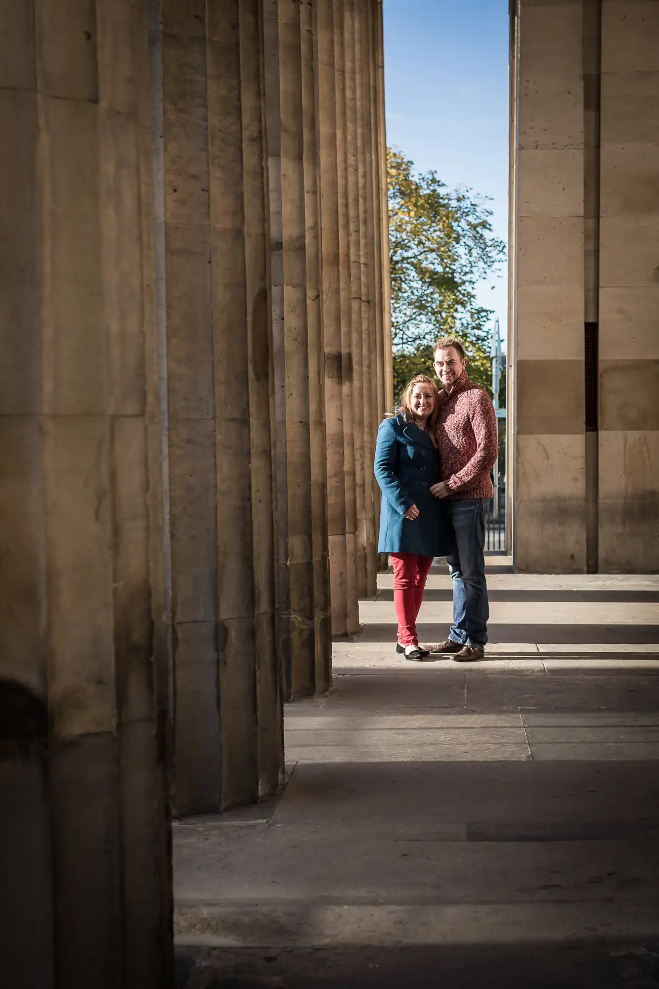 A couple embracing, standing between tall stone columns, with sunlight illuminating their smiling faces.