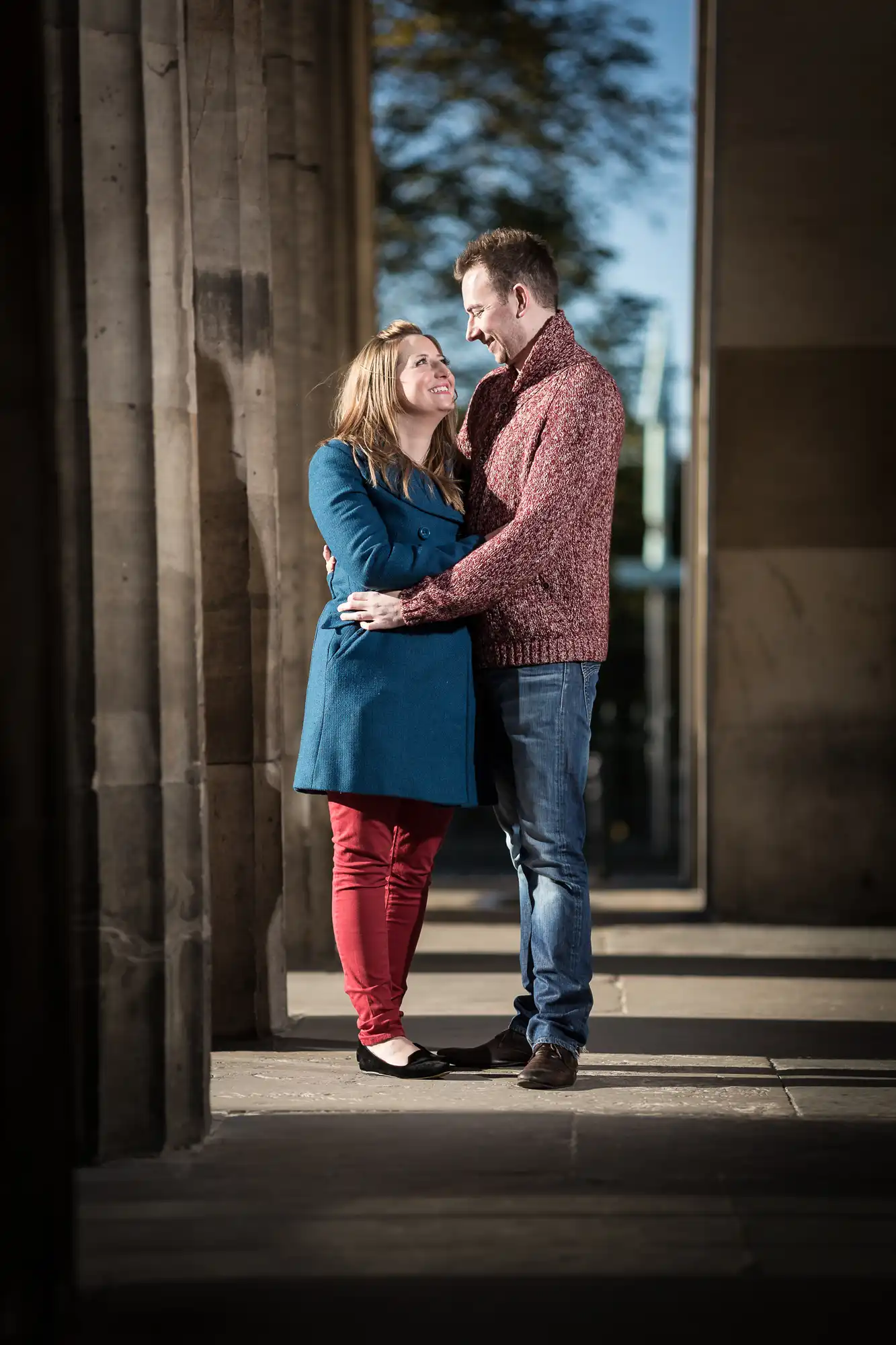 A couple embracing and smiling at each other between concrete columns, the woman in a blue coat and the man in a red sweater.