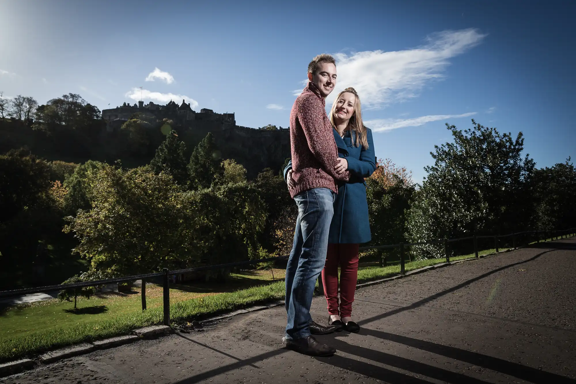 A couple embracing and smiling on a pathway, with a castle on a hill in the background and lush greenery under a bright sky.