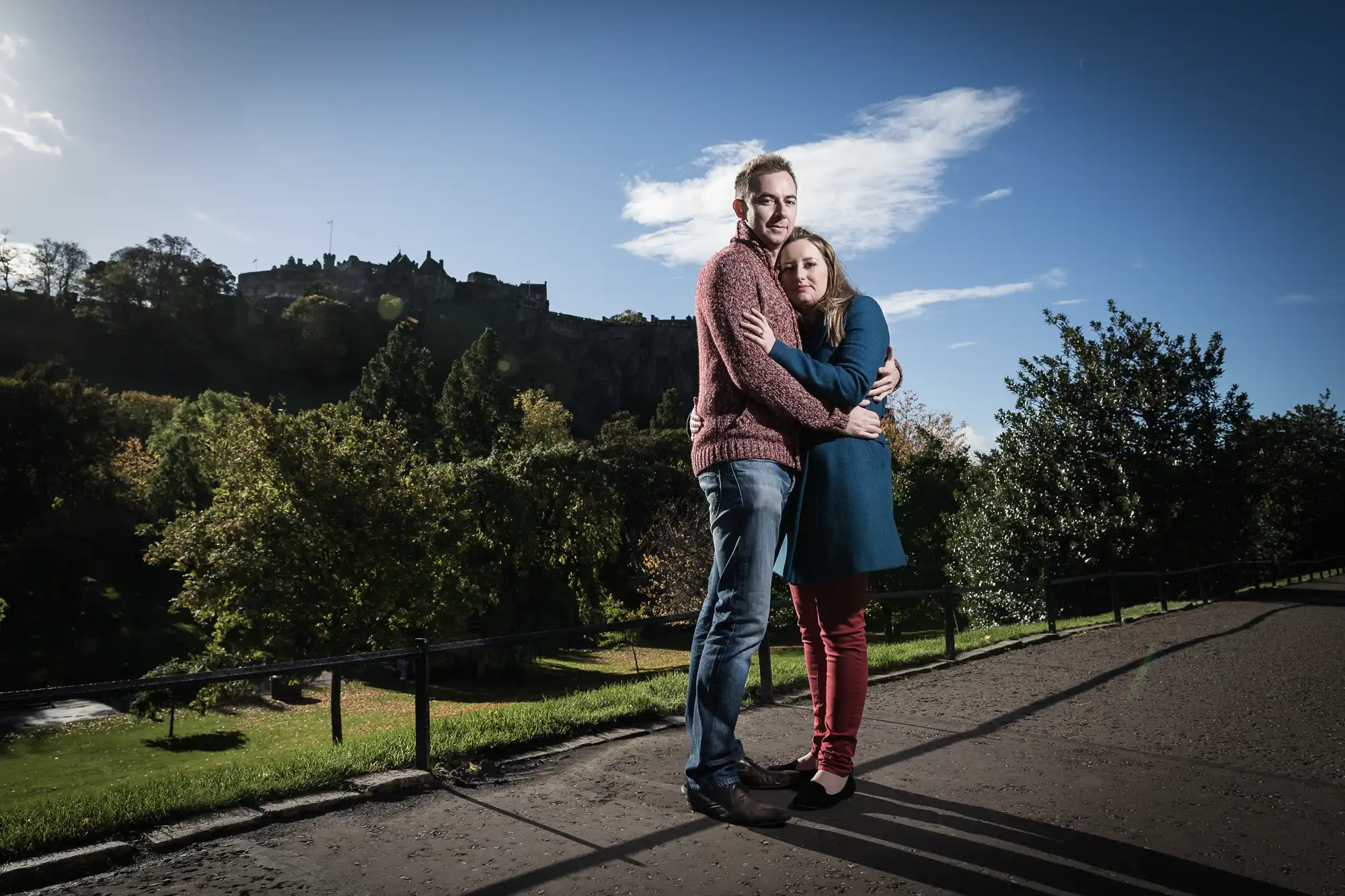 A couple embraces while standing on a path in a park, with a castle on a hill visible in the background under a blue sky.