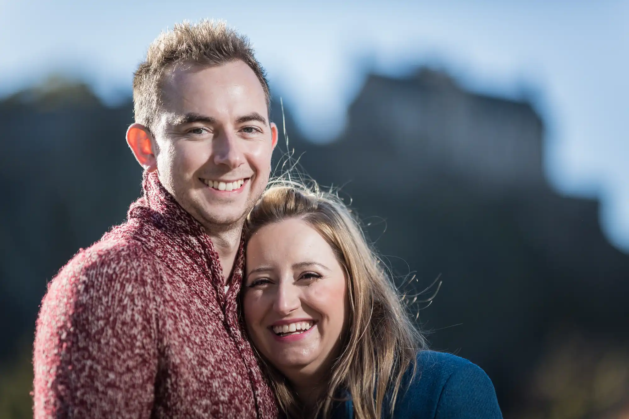 A smiling couple in warm clothing posing outdoors with a castle blurred in the background.