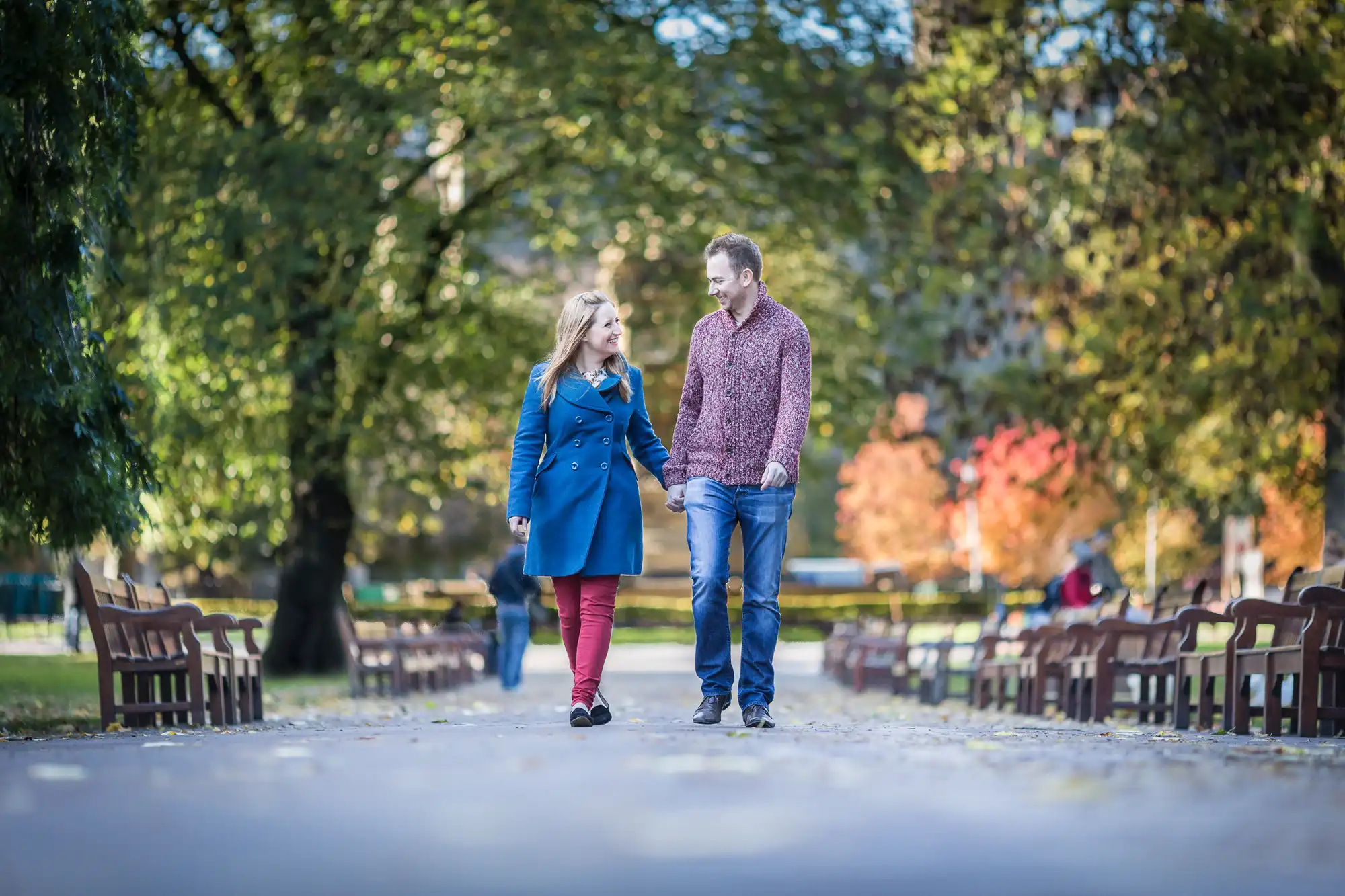 A couple walking hand in hand down a tree-lined pathway with fallen leaves and benches on either side, during autumn.