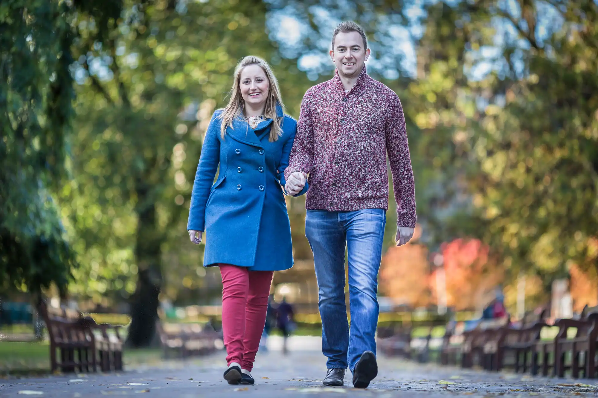 A couple holding hands and smiling while walking down a tree-lined park path in autumn.