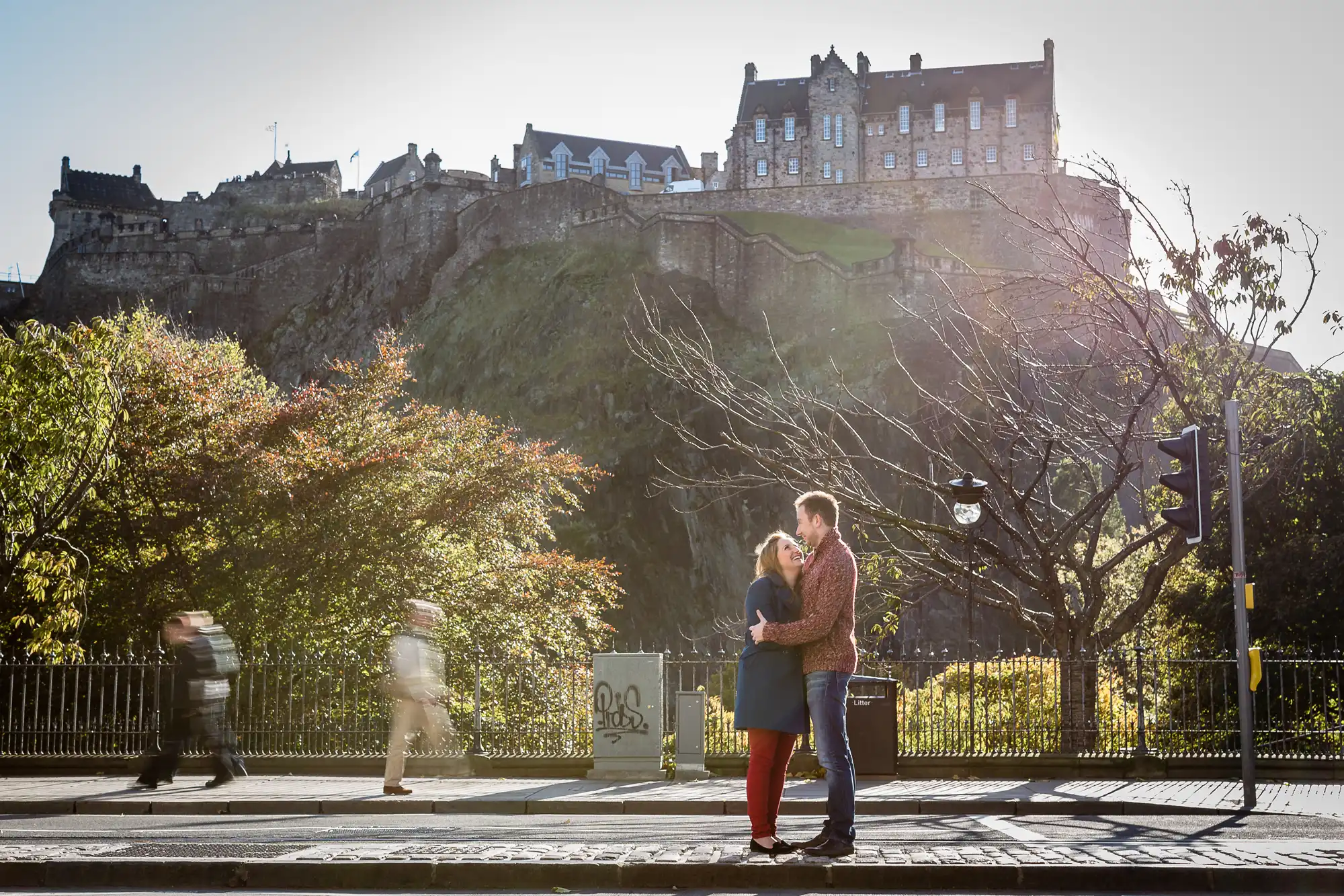 A couple embraces in the foreground with edinburgh castle perched atop a rocky hill in the background on a sunny day.