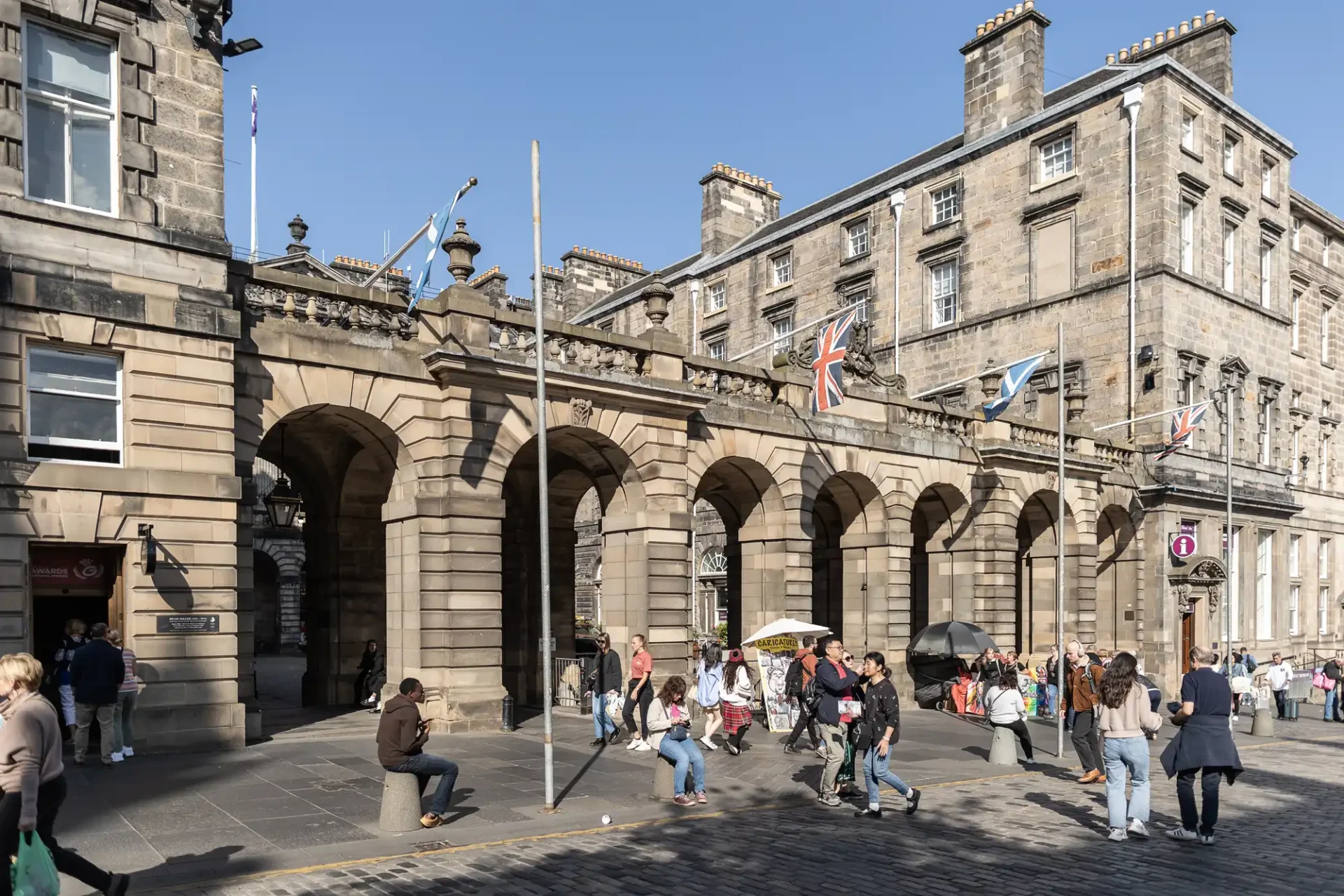 Historic stone building with arches and multiple flags in front, people walking and sitting nearby, under a clear blue sky.