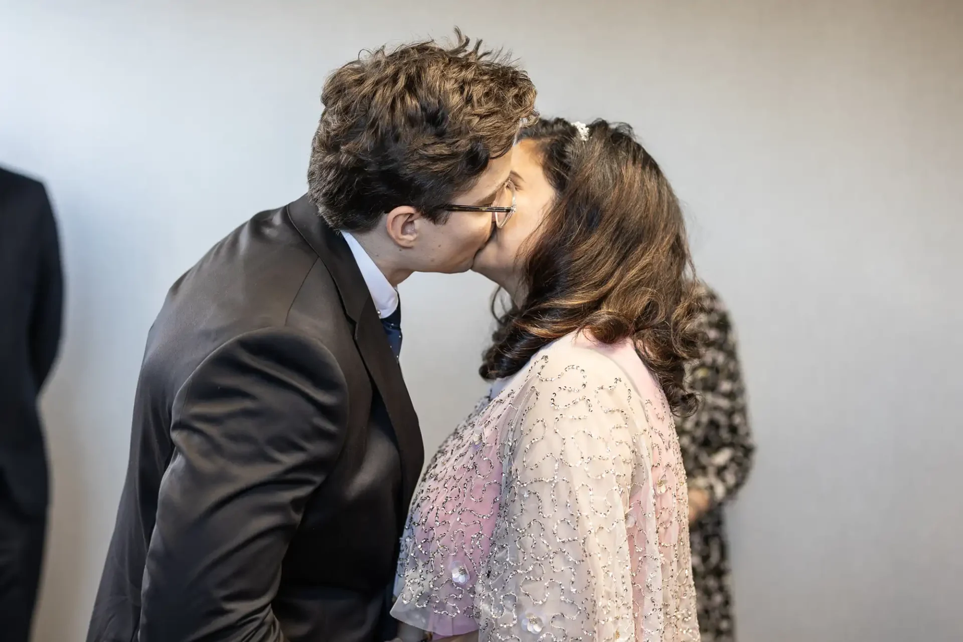 A couple in formal attire is kissing in a room where other people are present in the background.