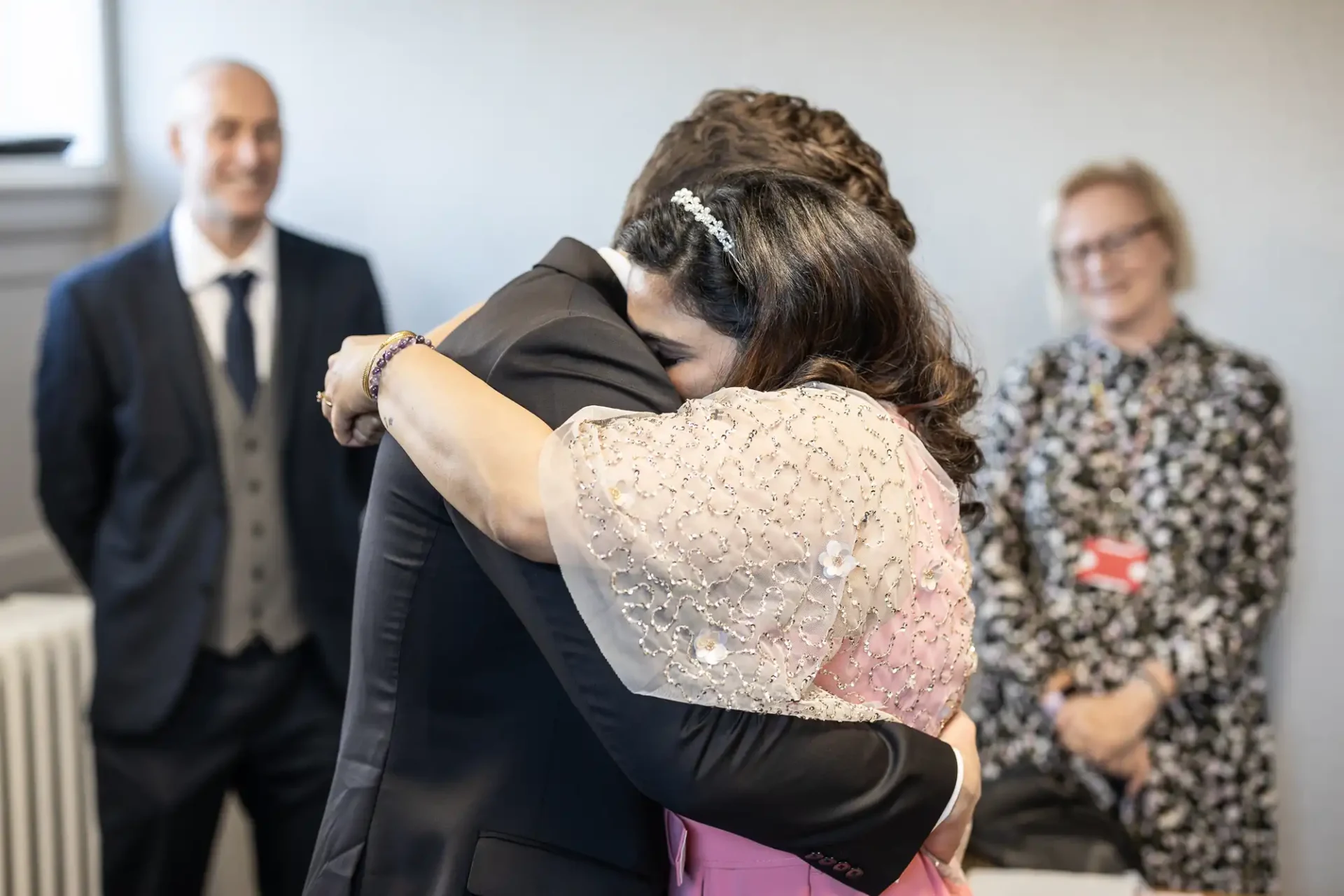 A woman in a pink dress and a man in a suit embrace in an indoor setting, while two people smile in the background.