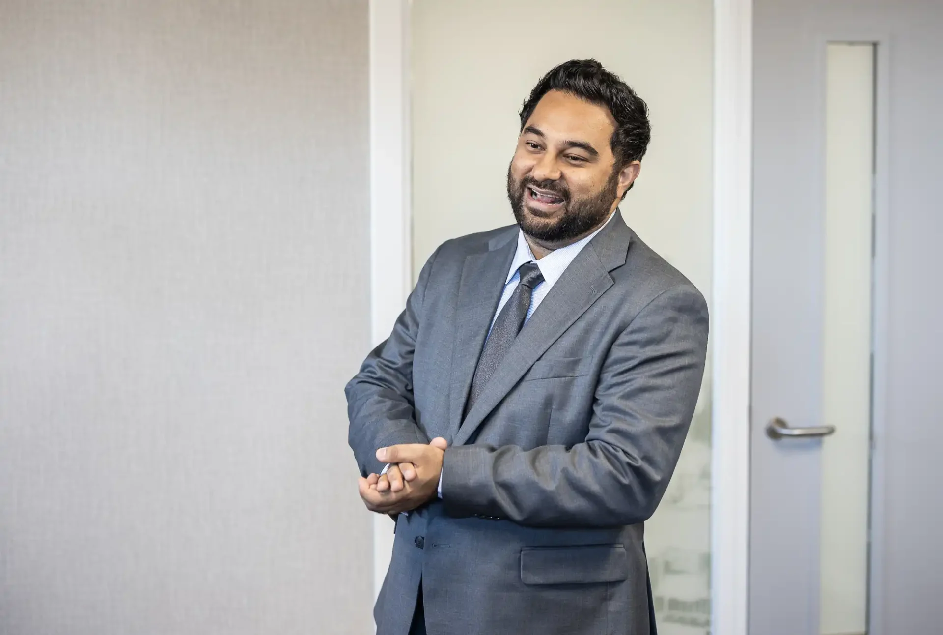 A man in a gray suit stands indoors, smiling with hands clasped.