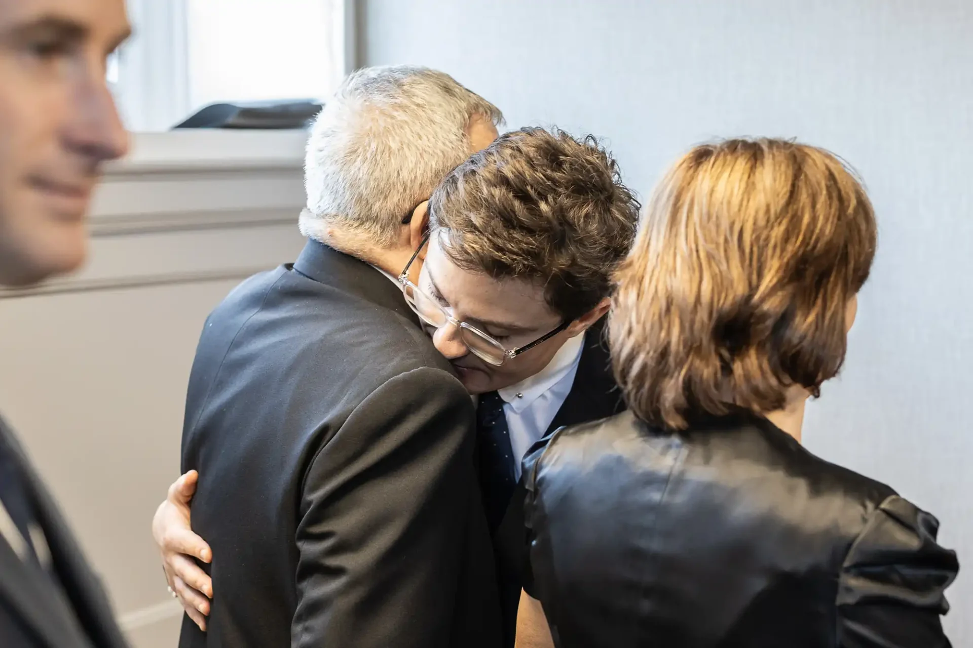 A group of people in a formal setting. Two men are hugging, while a woman with brown hair stands nearby with her back facing the camera.