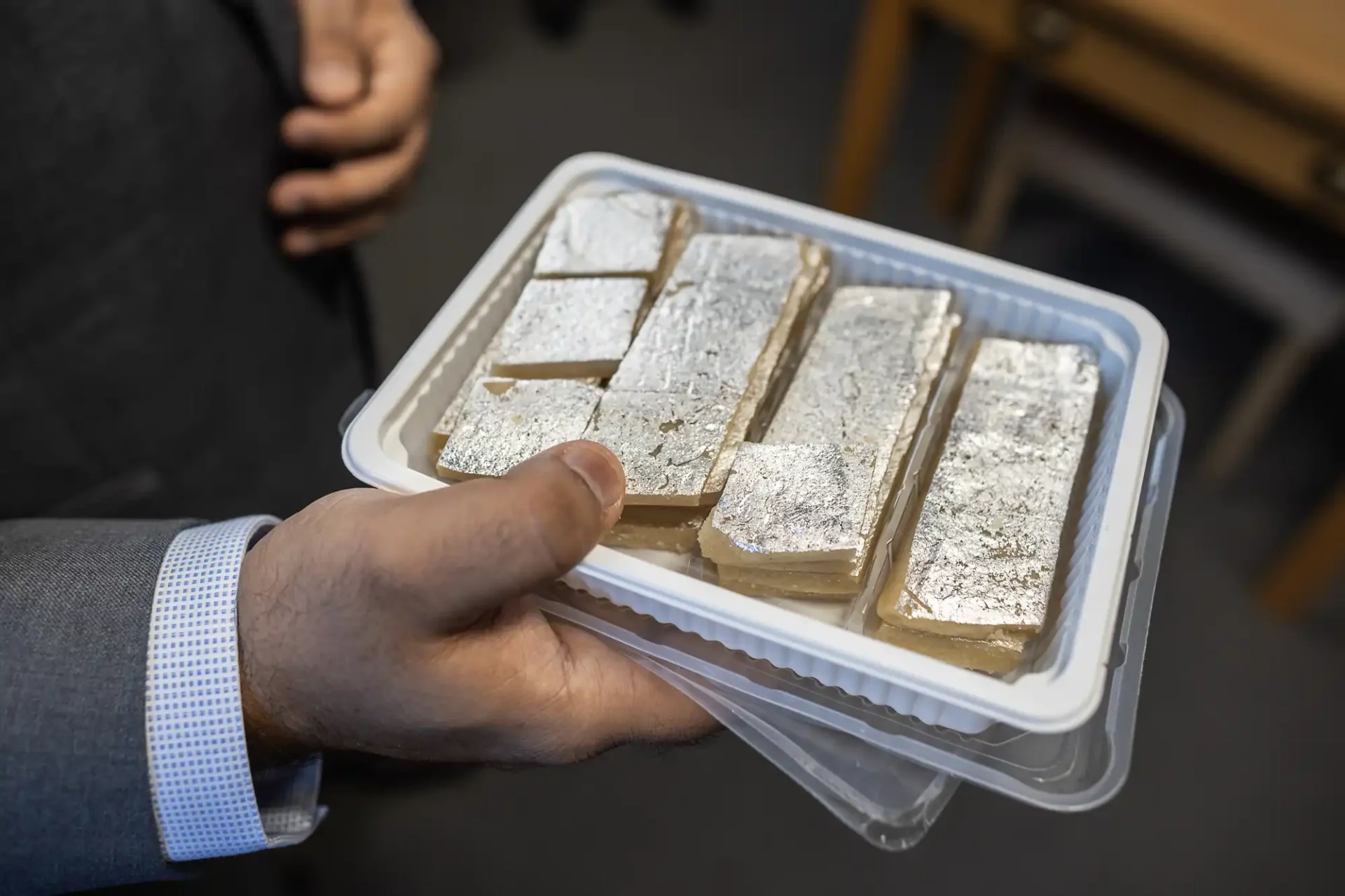 A person holds a plastic tray containing rectangular pieces of a dessert topped with edible silver foil.