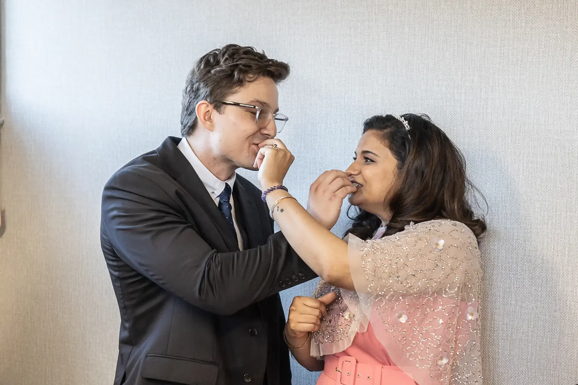 A man in a suit and a woman in a pink dress feeding each other cake in front of a light-colored wall.