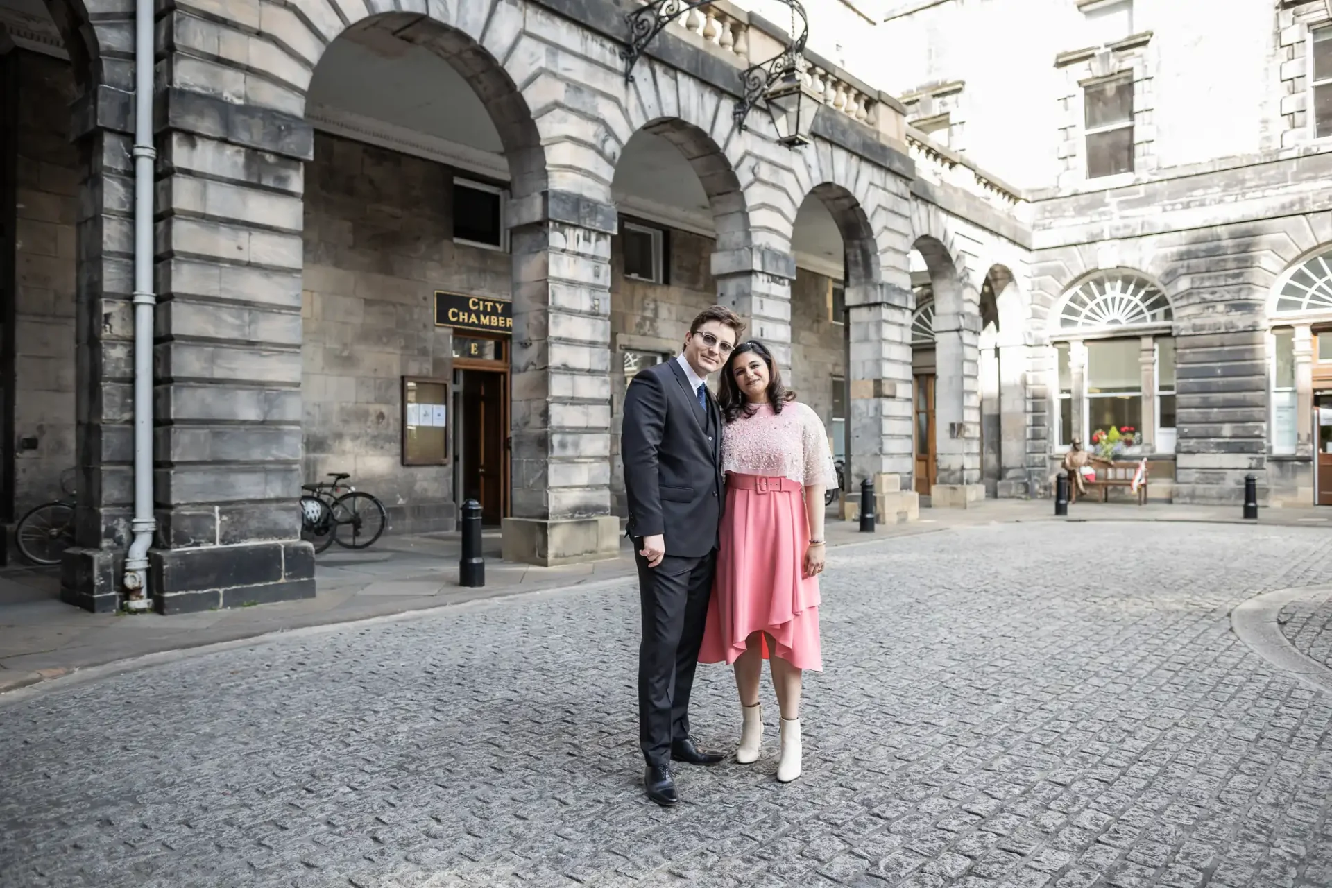 Two people stand together on a cobblestone street in front of a historic building with arches. They are dressed formally, with the woman in a pink dress and the man in a dark suit.