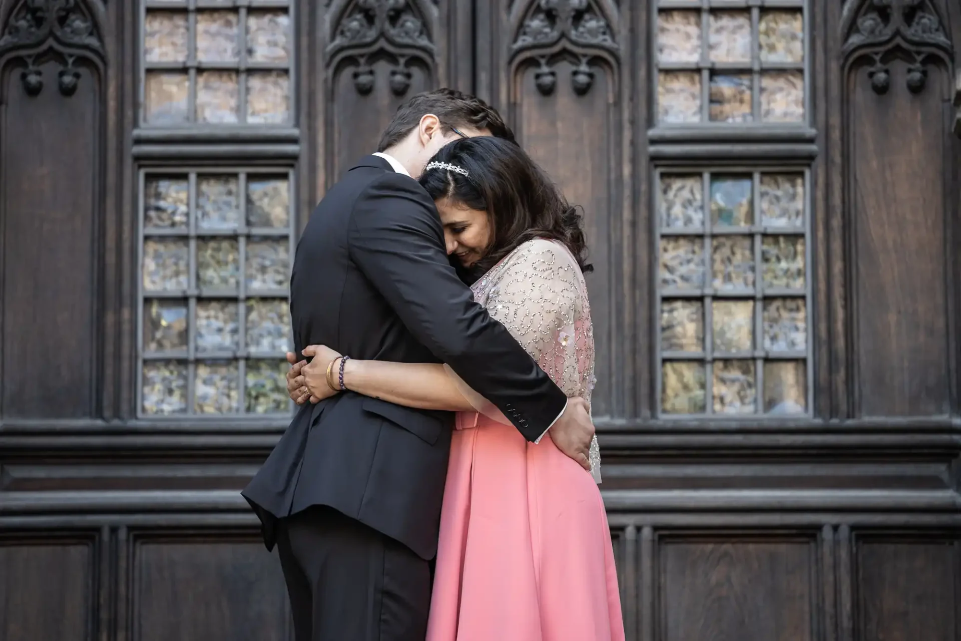 A couple embraces in front of an intricate wooden door with stained glass windows. The woman wears a pink dress, and the man is in a black suit.