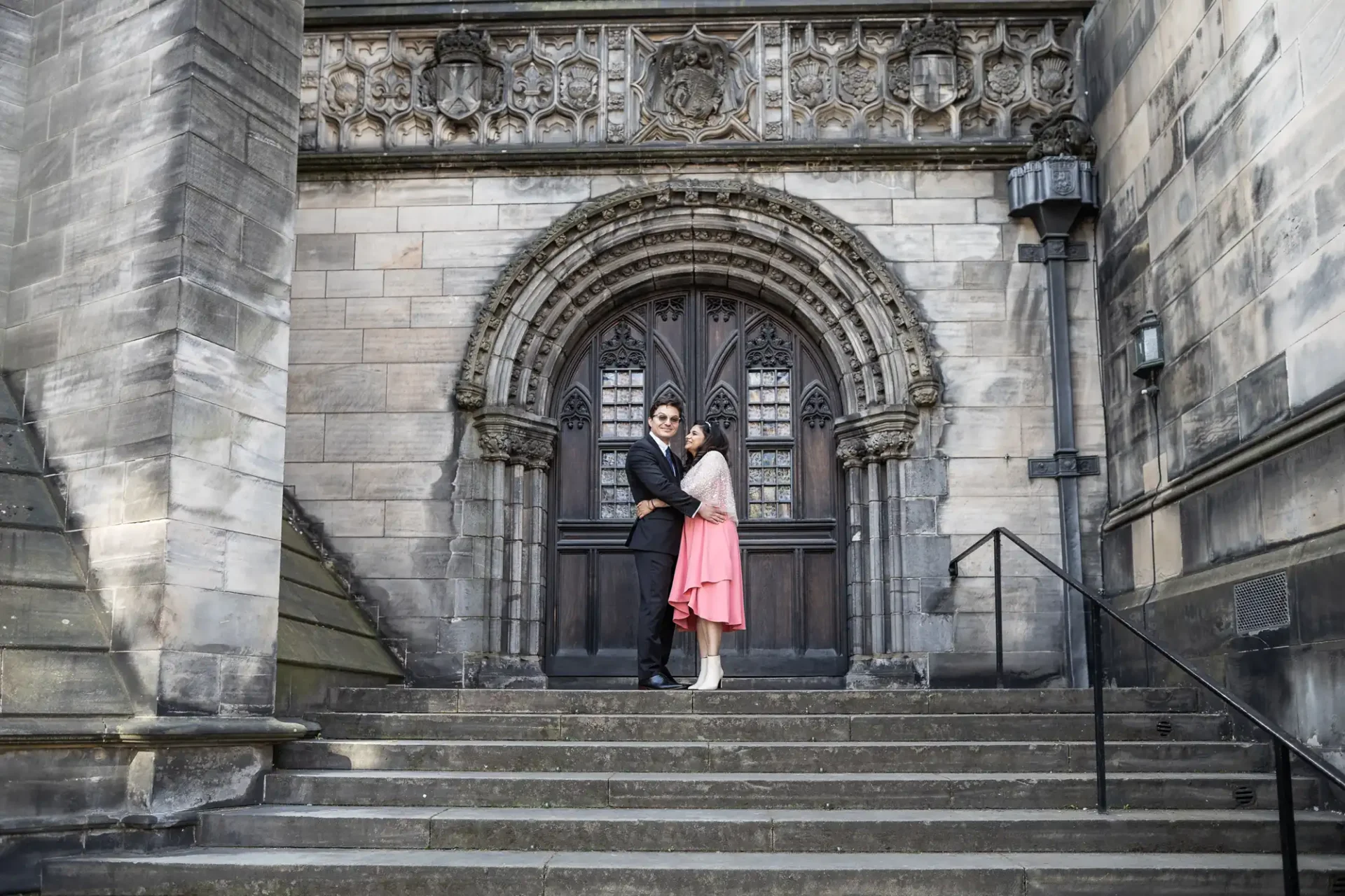 A couple poses on stone steps in front of an ornate, arched wooden door set in a Gothic-style stone building. The man wears a black suit, and the woman is in a pink dress.