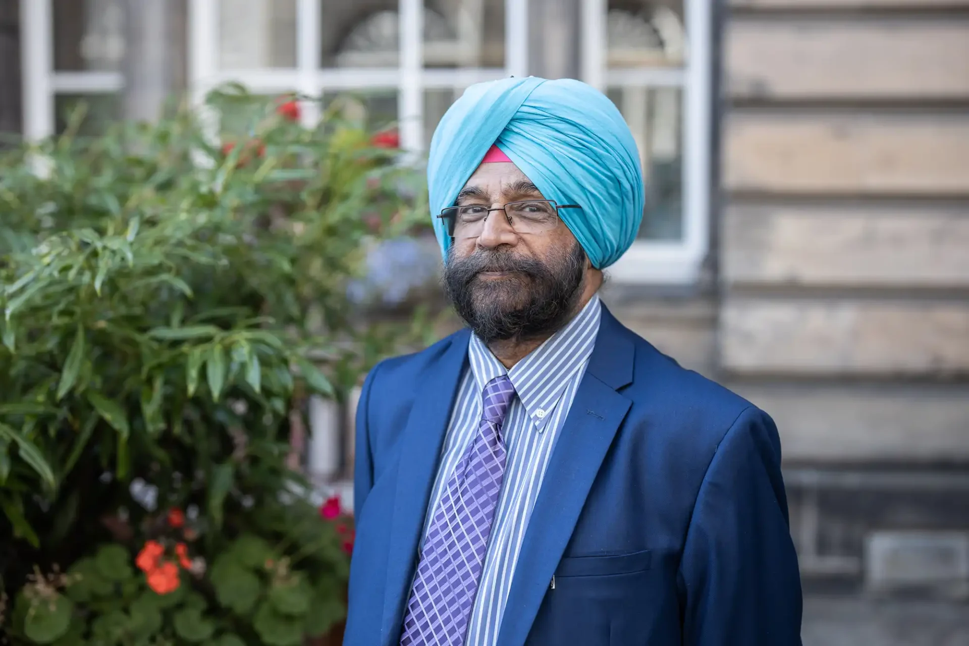 A man wearing a blue turban, glasses, and a blue suit with a patterned tie stands outdoors near a bush with flowers.