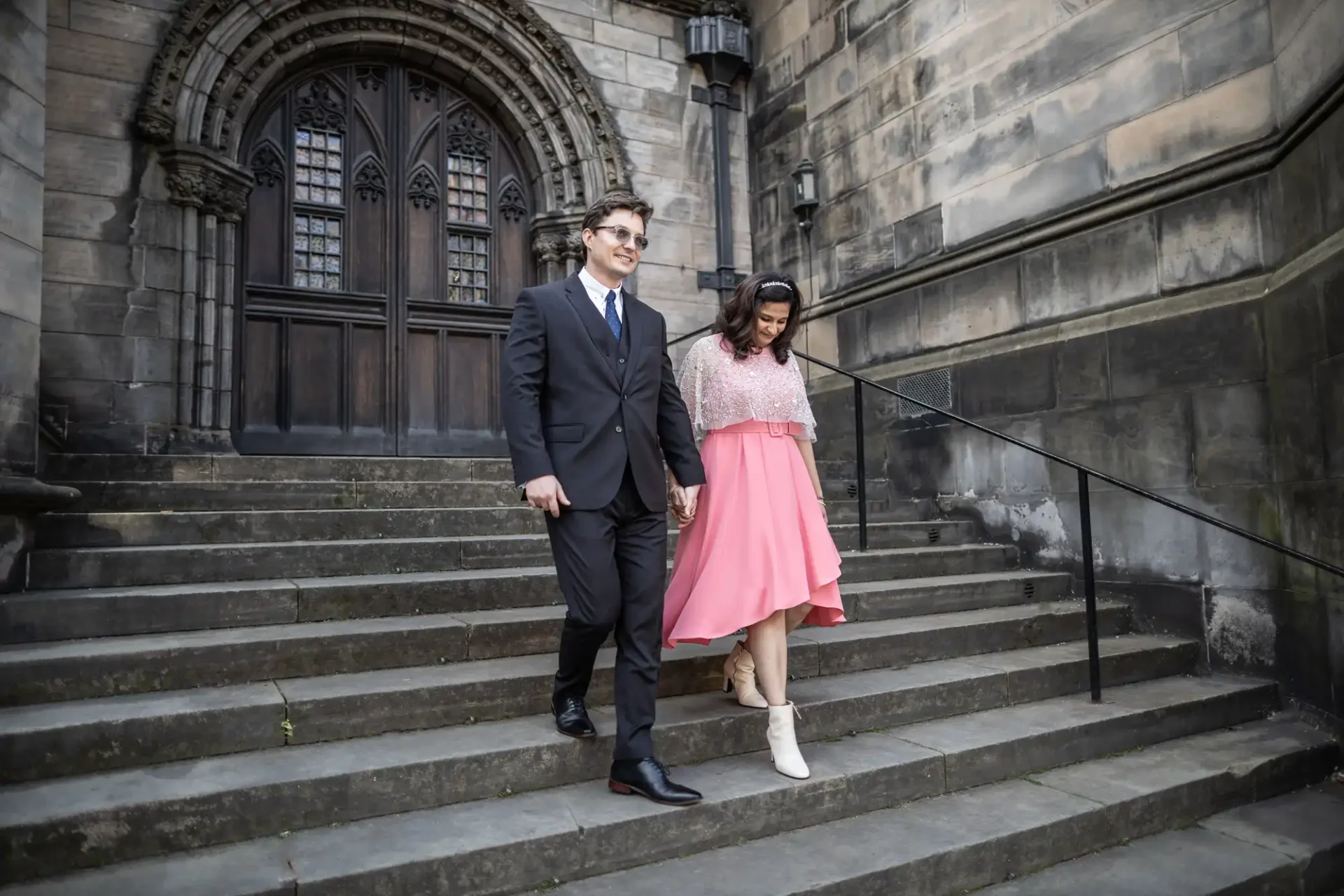 A man in a suit and a woman in a pink dress walk down stone steps outside a historic building.