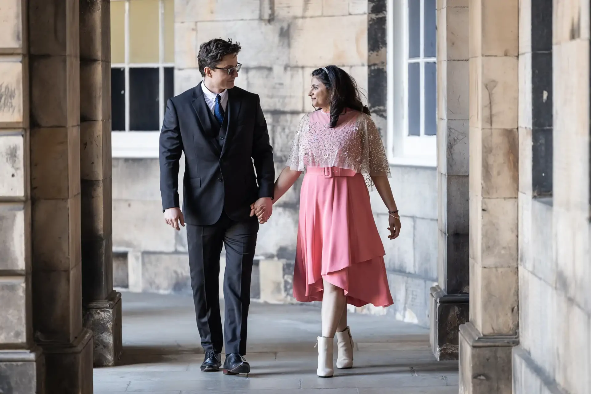 A man in a suit and a woman in a pink dress hold hands while walking in a corridor with stone walls and arched openings.