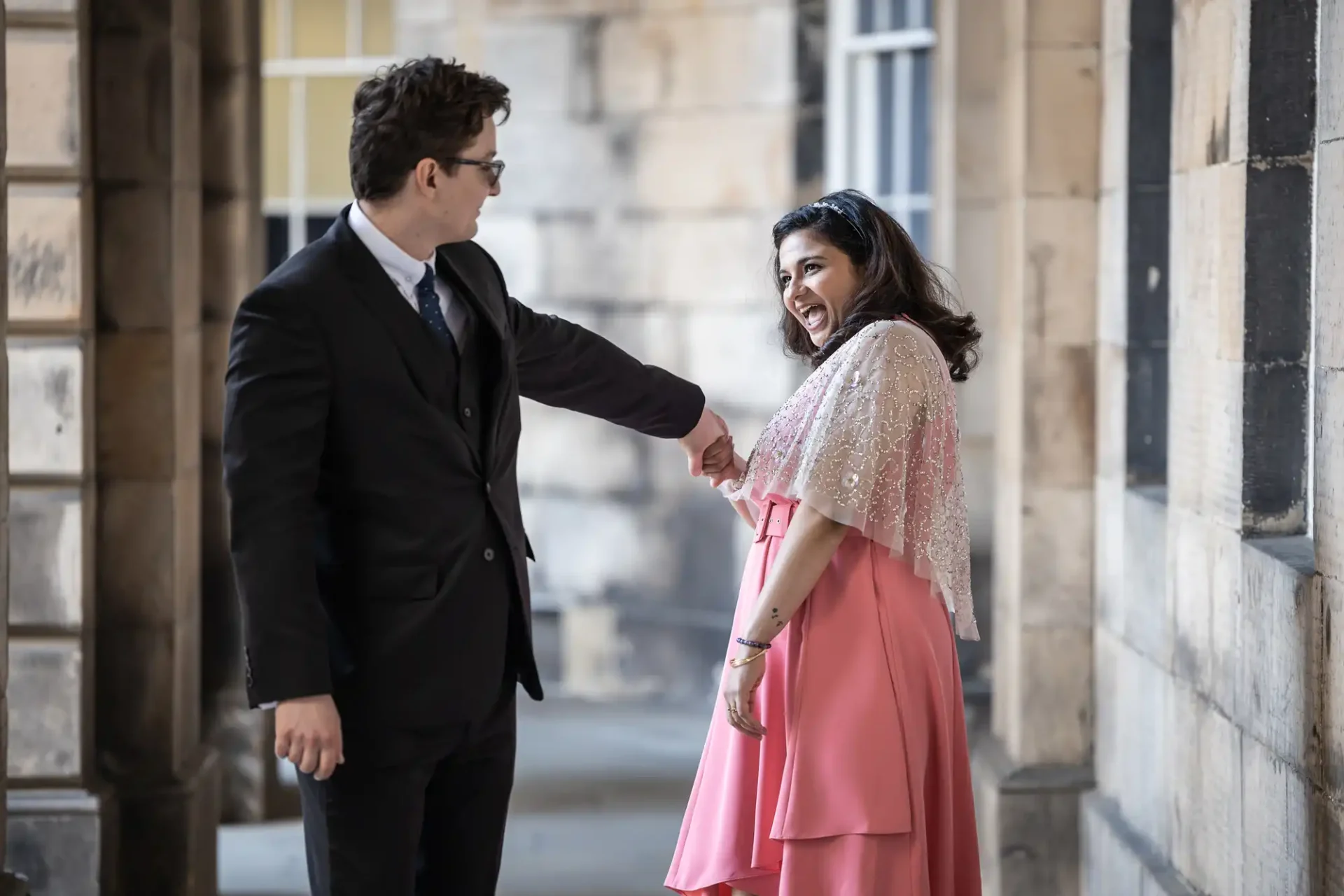 A man in a suit holds hands with a woman in a pink dress and lace shawl, smiling, standing in a stone corridor.