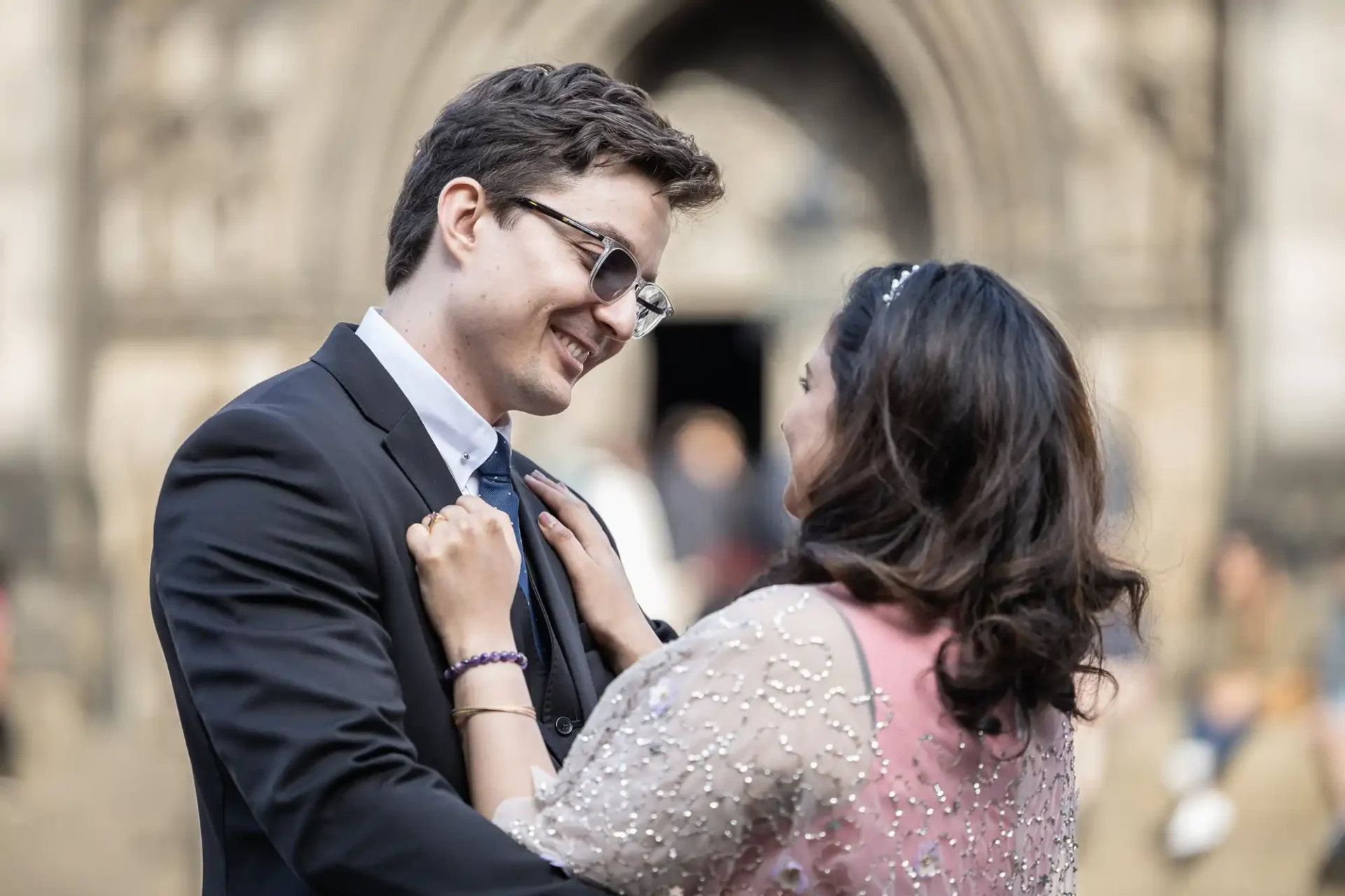 A couple smiles while standing close, facing each other. The man wears a suit and glasses; the woman, a dress with a sheer embellished overlay. A Gothic-style building is in the background.