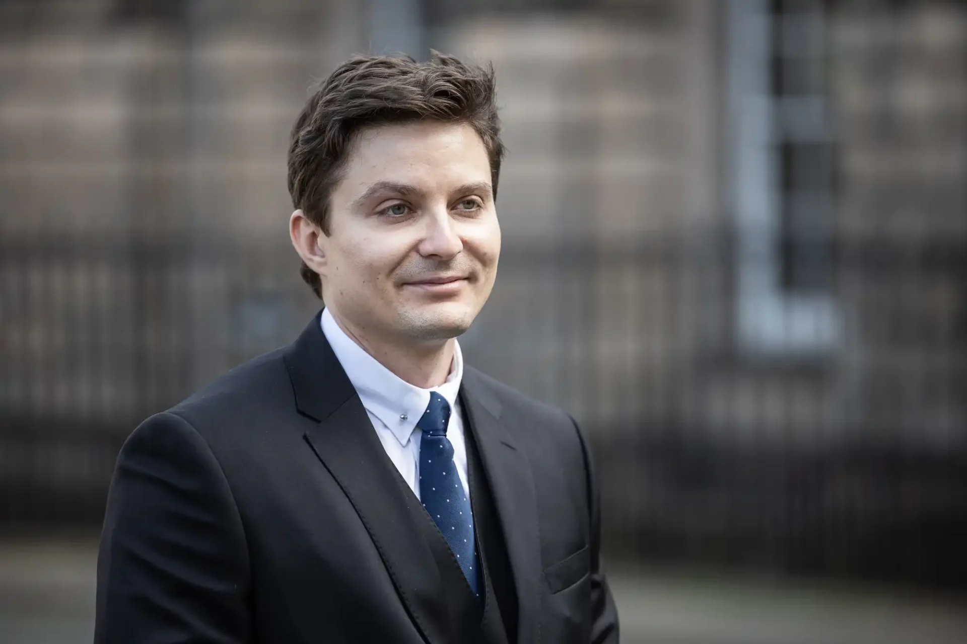 A man in a dark suit and blue tie with a white shirt stands outside, looking forward. The background is blurred, showing a building facade.