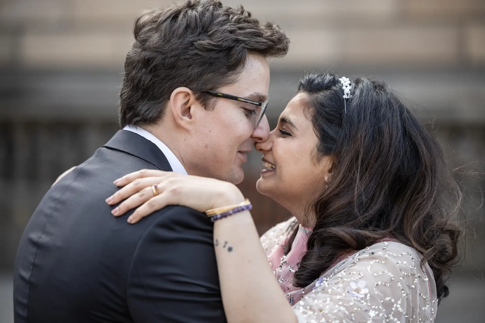 A couple embraces, smiling and touching noses. The woman wears a tiara and embellished attire, while the man is in a dark suit.