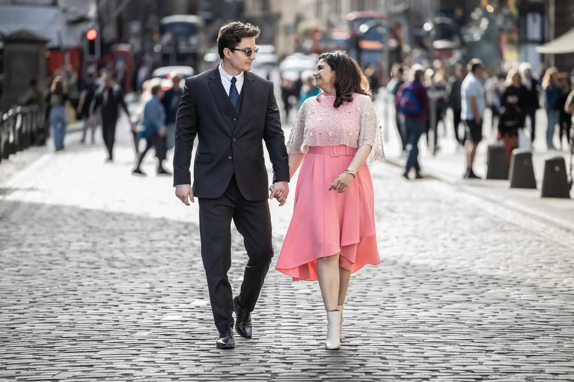 A man in a suit and tie and a woman in a pink dress hold hands while walking on a cobblestone street in a busy city.