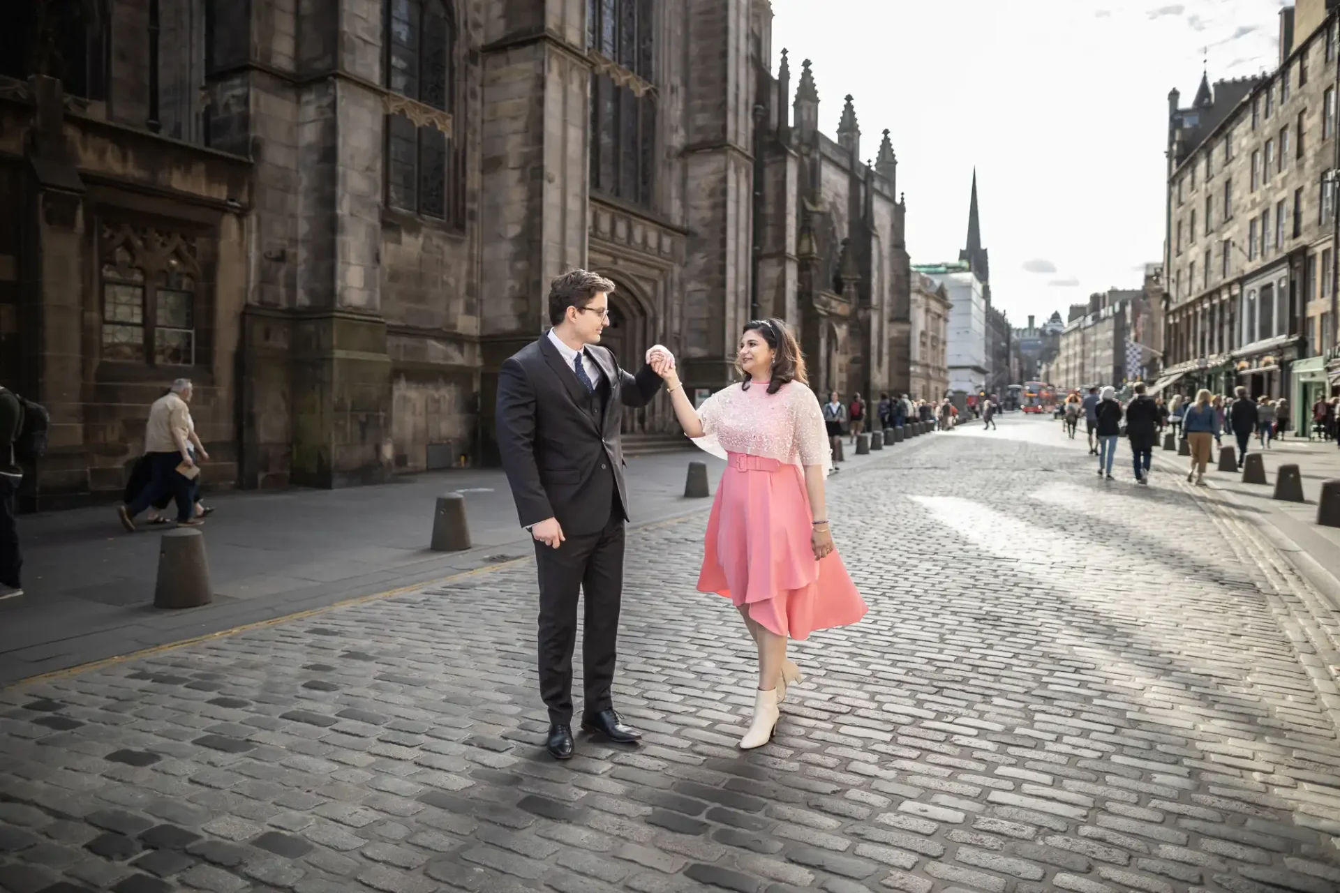 A man in a suit and a woman in a pink dress hold hands on a cobblestone street, with historic buildings in the background.