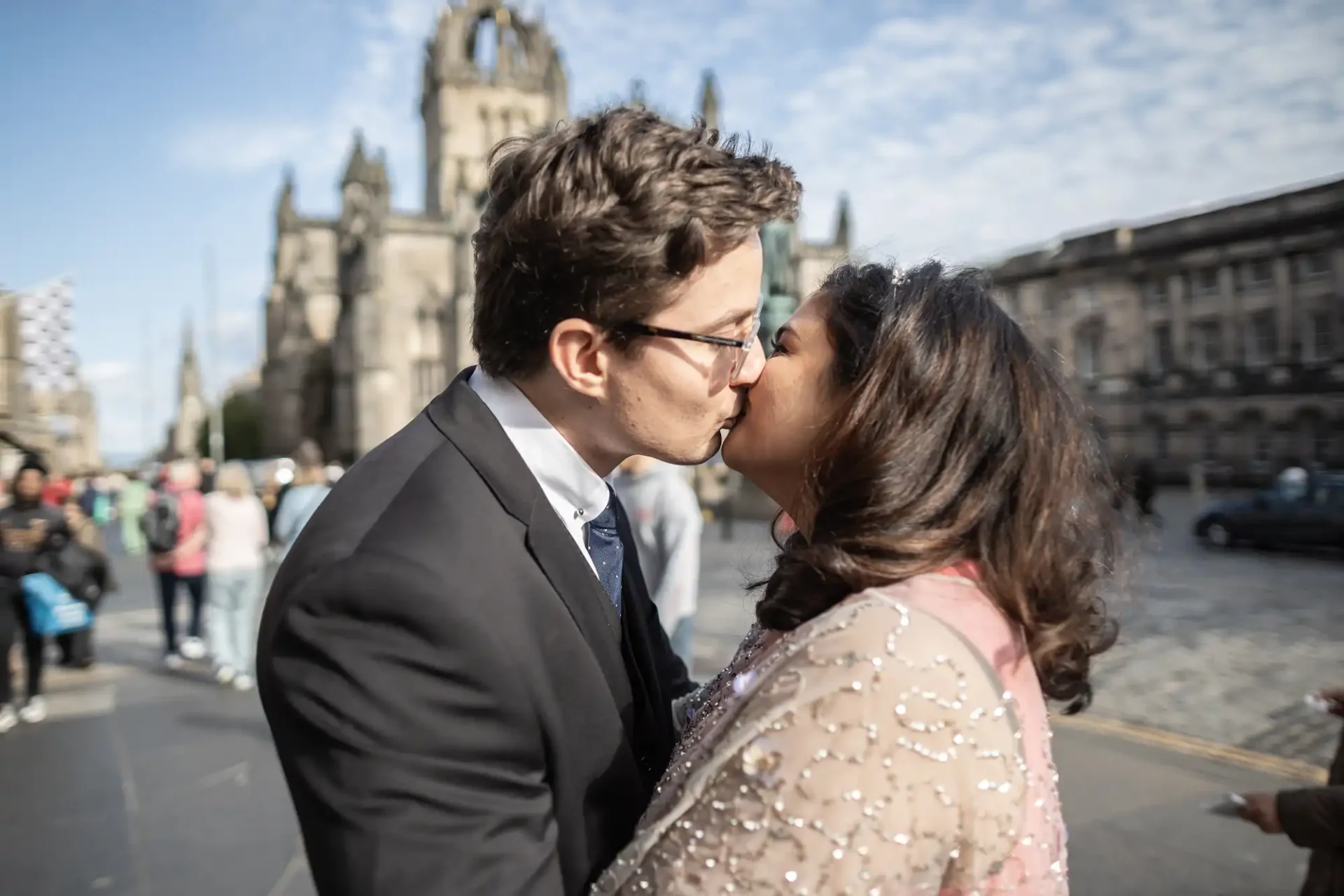 A couple kisses in front of a historic church on a busy street, surrounded by people.