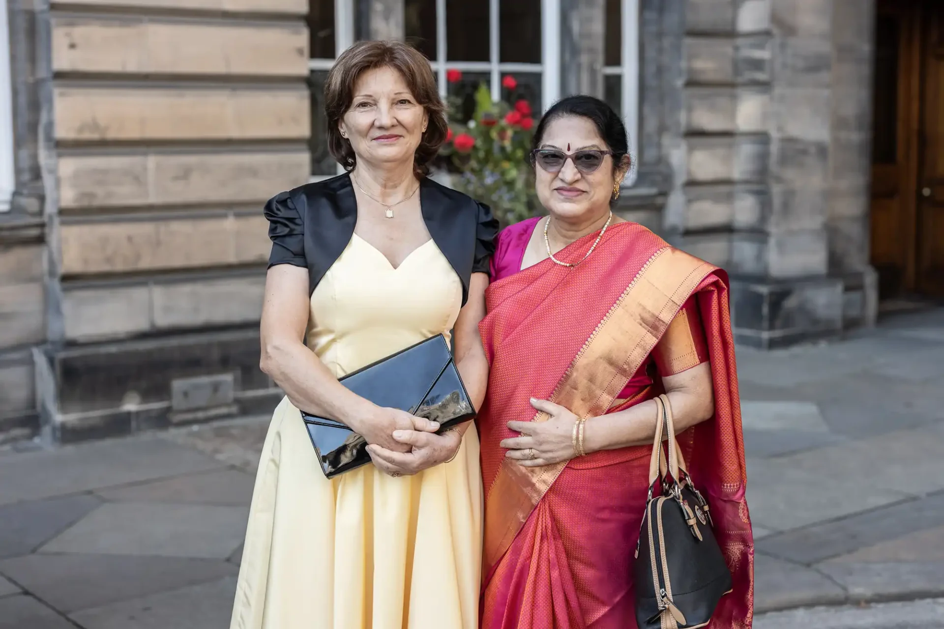 Two women standing outdoors, one in a yellow dress holding a clutch, the other in a red and gold sari, both smiling. Stone building and flowering plant in the background.