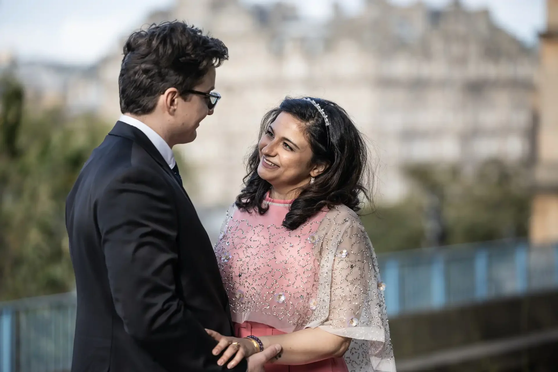 A couple stands outdoors, smiling at each other. The man wears a suit and glasses, while the woman is in a pink dress with an embellished sheer overlay. A blurred building is in the background.