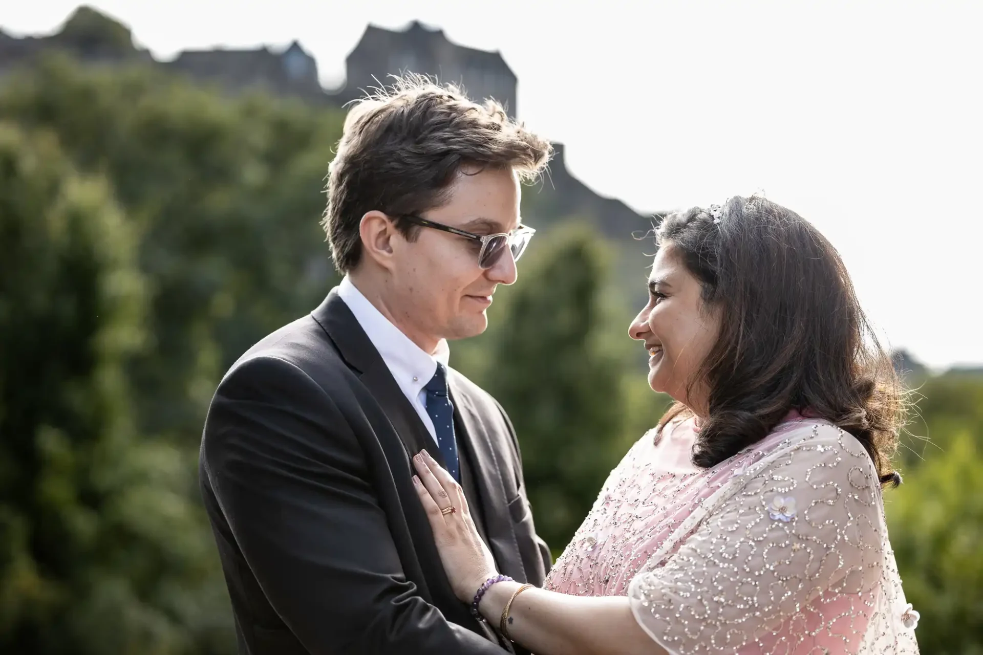 A man and woman in formal attire stand close together outdoors, smiling at each other.