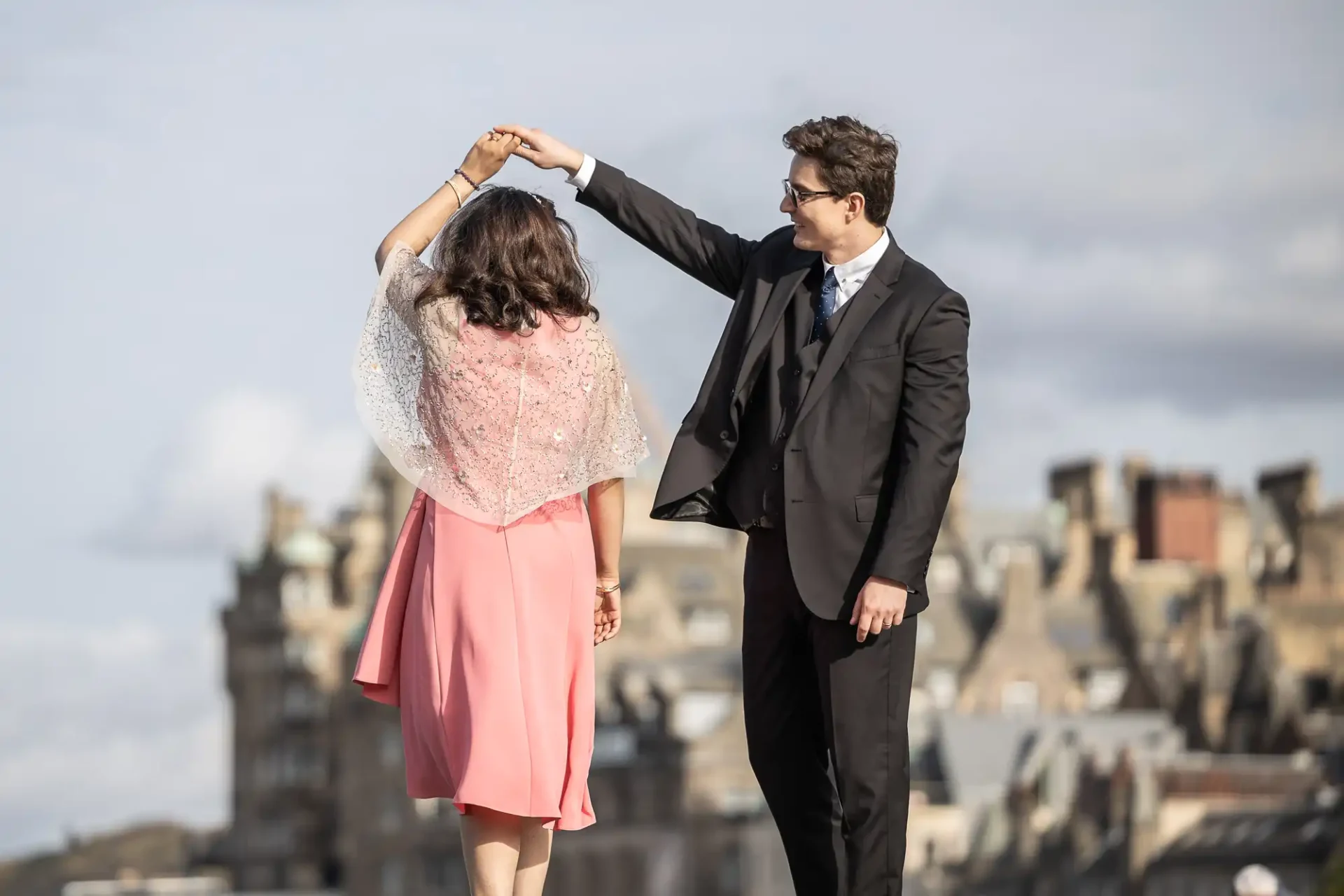 A man in a suit twirls a woman in a pink dress outdoors, with historical buildings in the background.