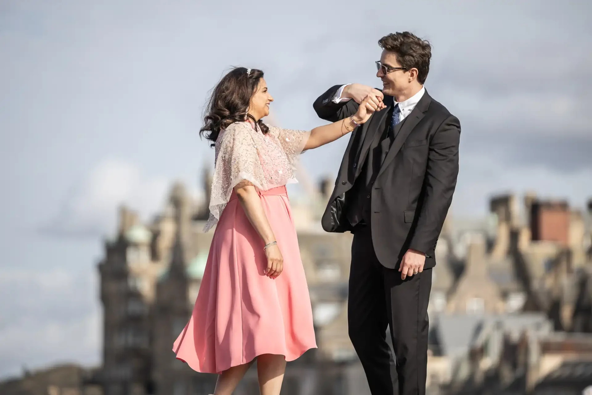 A woman in a pink dress holds hands and laughs with a man in a suit against a backdrop of blurred historic buildings.