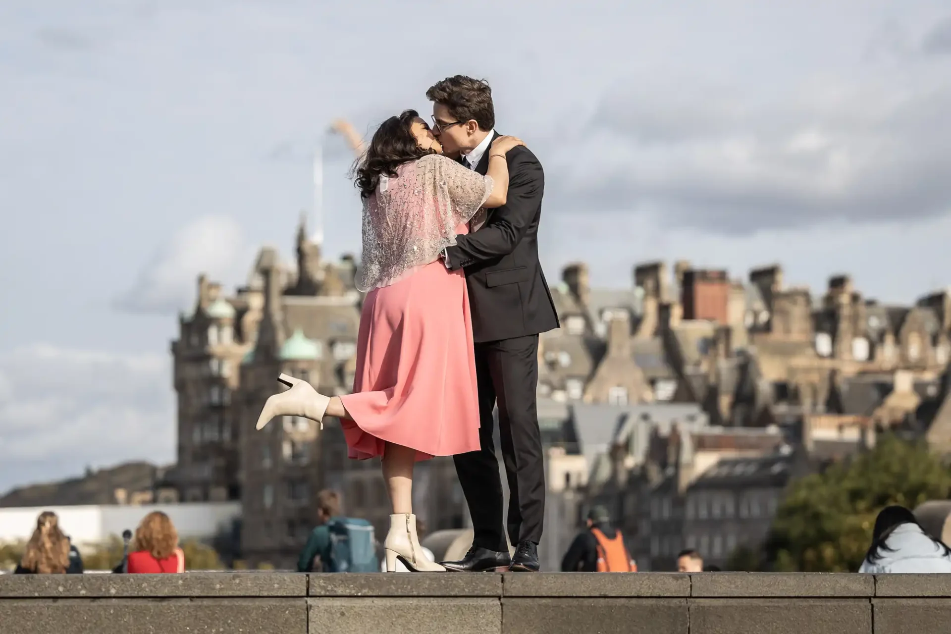 A couple in formal attire kisses on a stone ledge, with historic buildings in the background. The woman is wearing a pink dress and white boots, and the man is in a black suit.