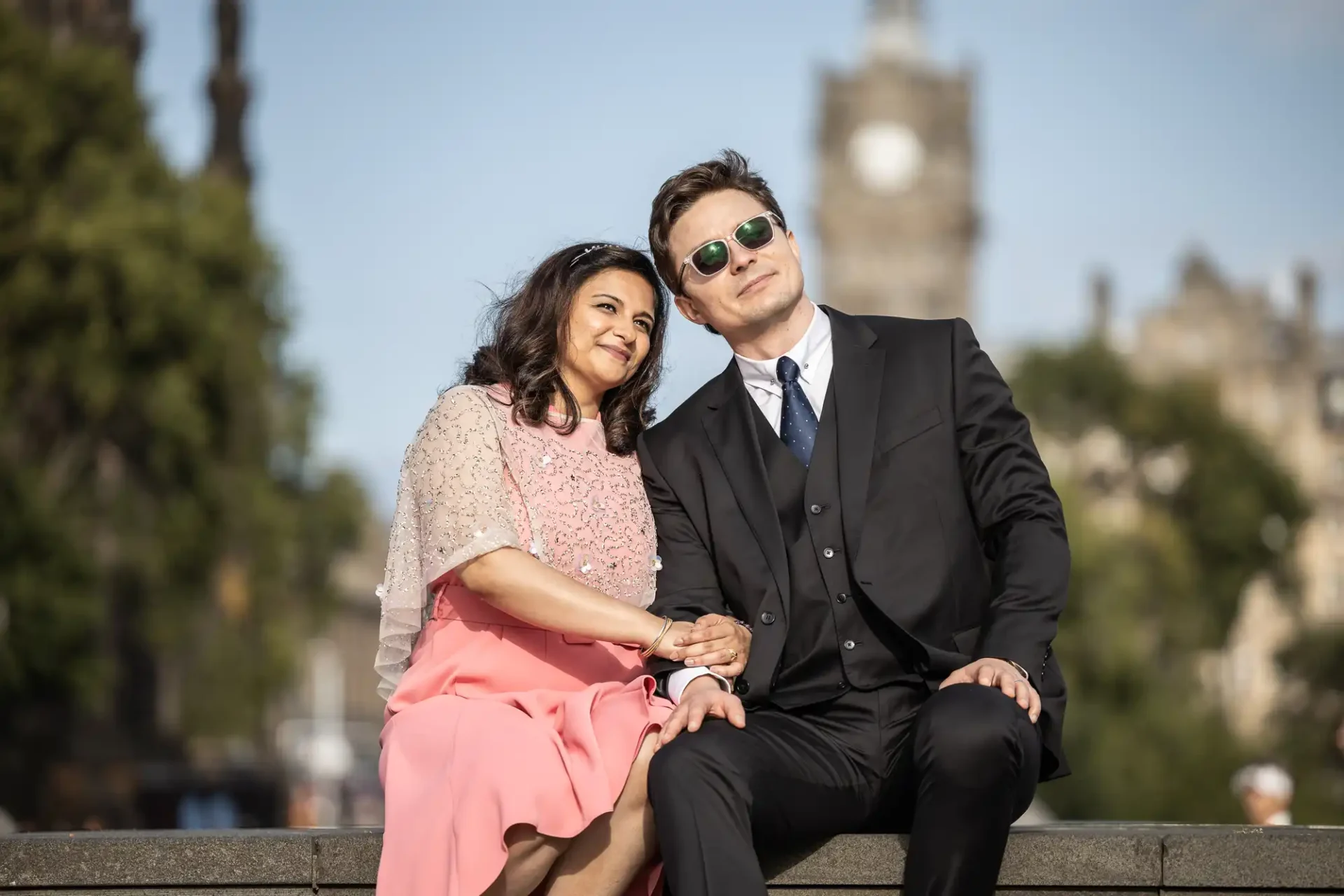 A woman in a pink dress and a man in a black suit sit together outdoors, holding hands and looking up, with a blurred cityscape in the background.