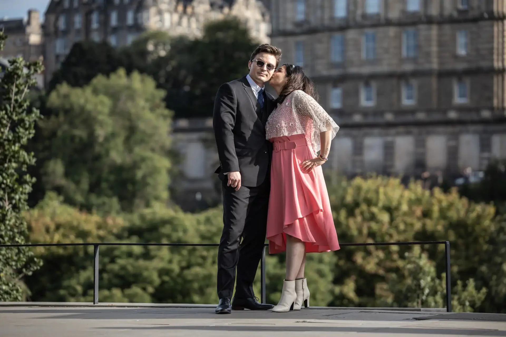 A man in a suit and sunglasses stands next to a woman in a pink dress kissing his cheek. They are outdoors, with a large building and trees in the background.