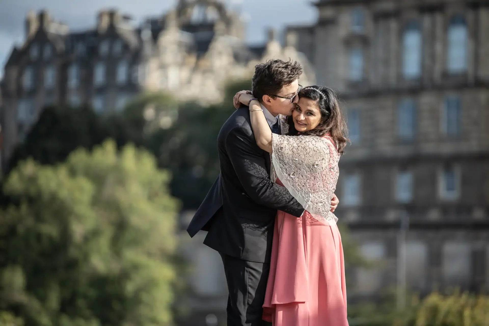 A couple embraces outdoors; the man in a black suit kisses the woman, who is wearing a pink dress and lace shawl. A historic building and trees are in the background.