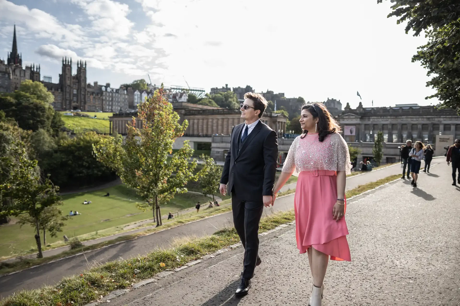 A couple walks hand in hand on a pathway in a park, with trees and historic buildings in the background.