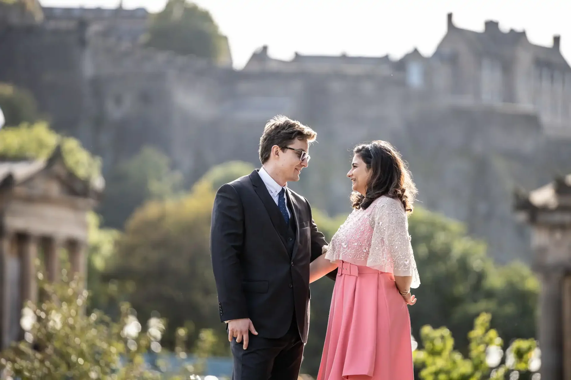 A couple stands outdoors, facing each other and smiling. They are dressed formally. A large, blurred stone building is visible in the background.