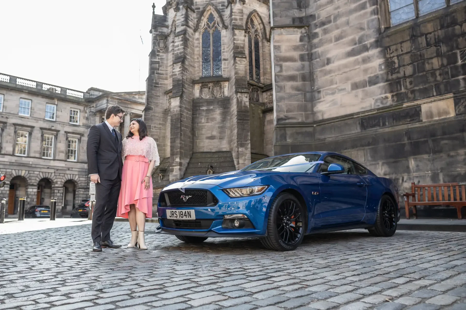 A man and woman stand on a cobblestone street next to a blue Ford Mustang. A historic building is in the background.