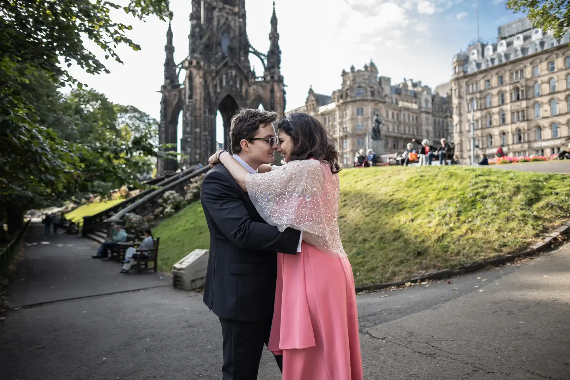 A couple embraces in a park near a Gothic-style monument and historic buildings. The woman wears a pink dress and the man is in a black suit.