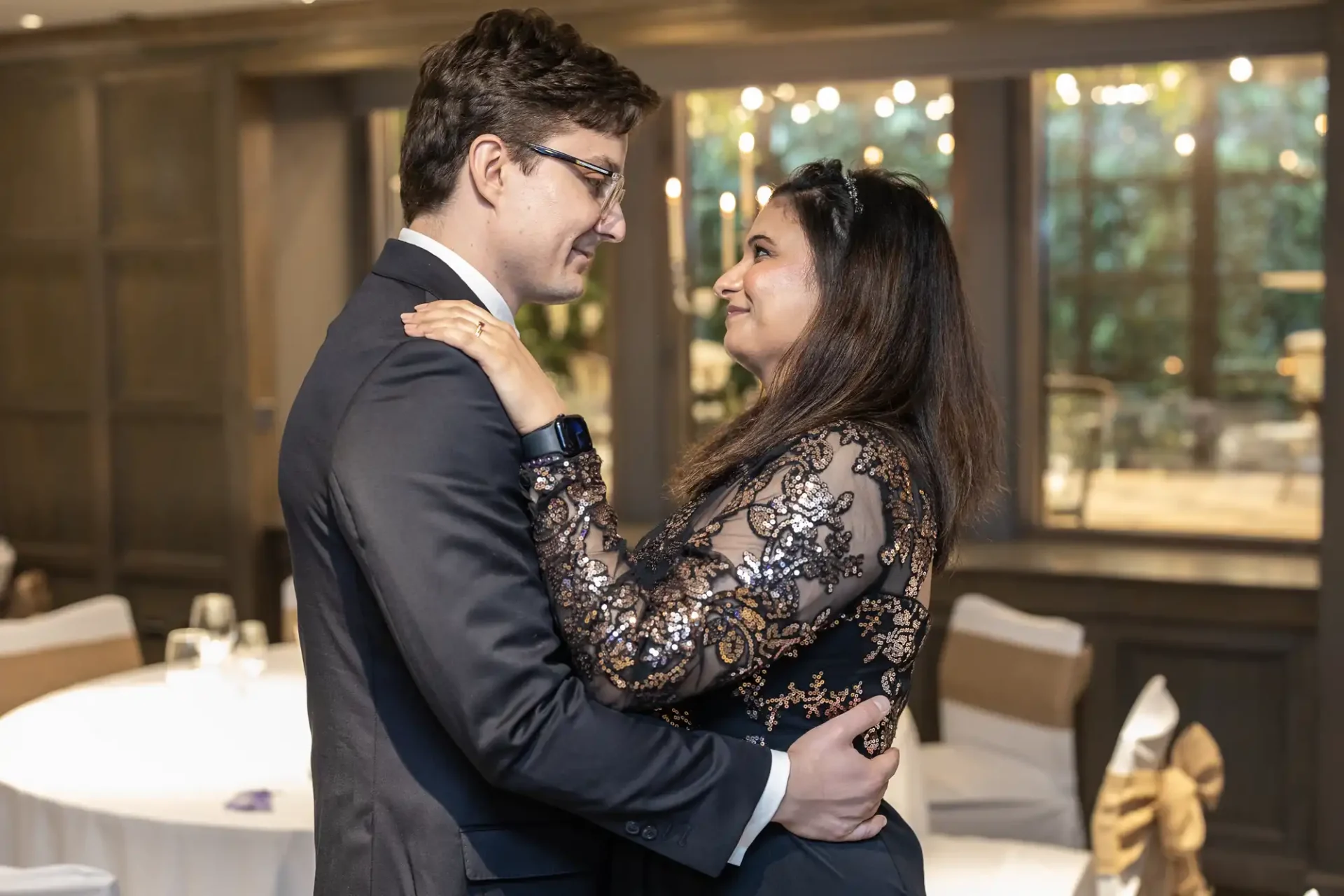 Two people dressed formally are standing closely, facing each other, with their hands touching. They are indoors, with round tables and chairs visible in the background.