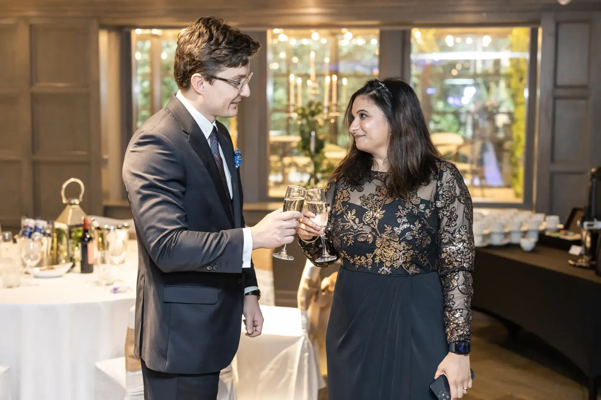 A man and woman in formal attire clink champagne glasses at an indoor event with tables, a gift bag, and decorations in the background.